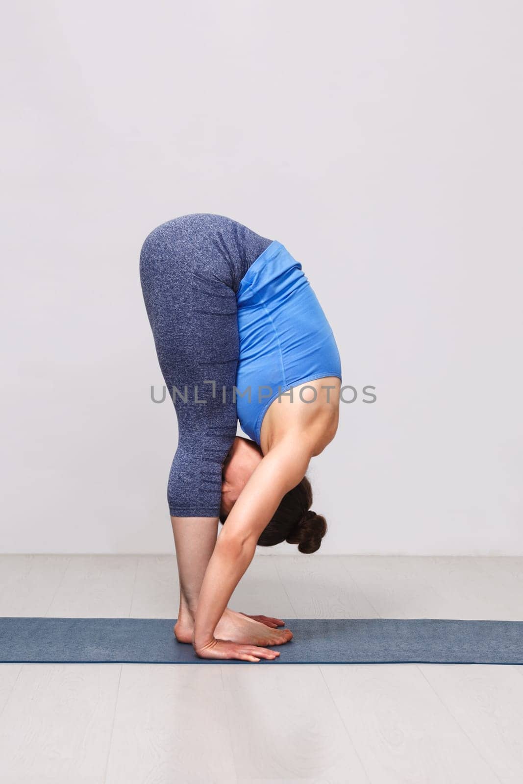Woman doing yoga asana Uttanasana - standing forward bend pose on yoga mat in studio on grey bagckground