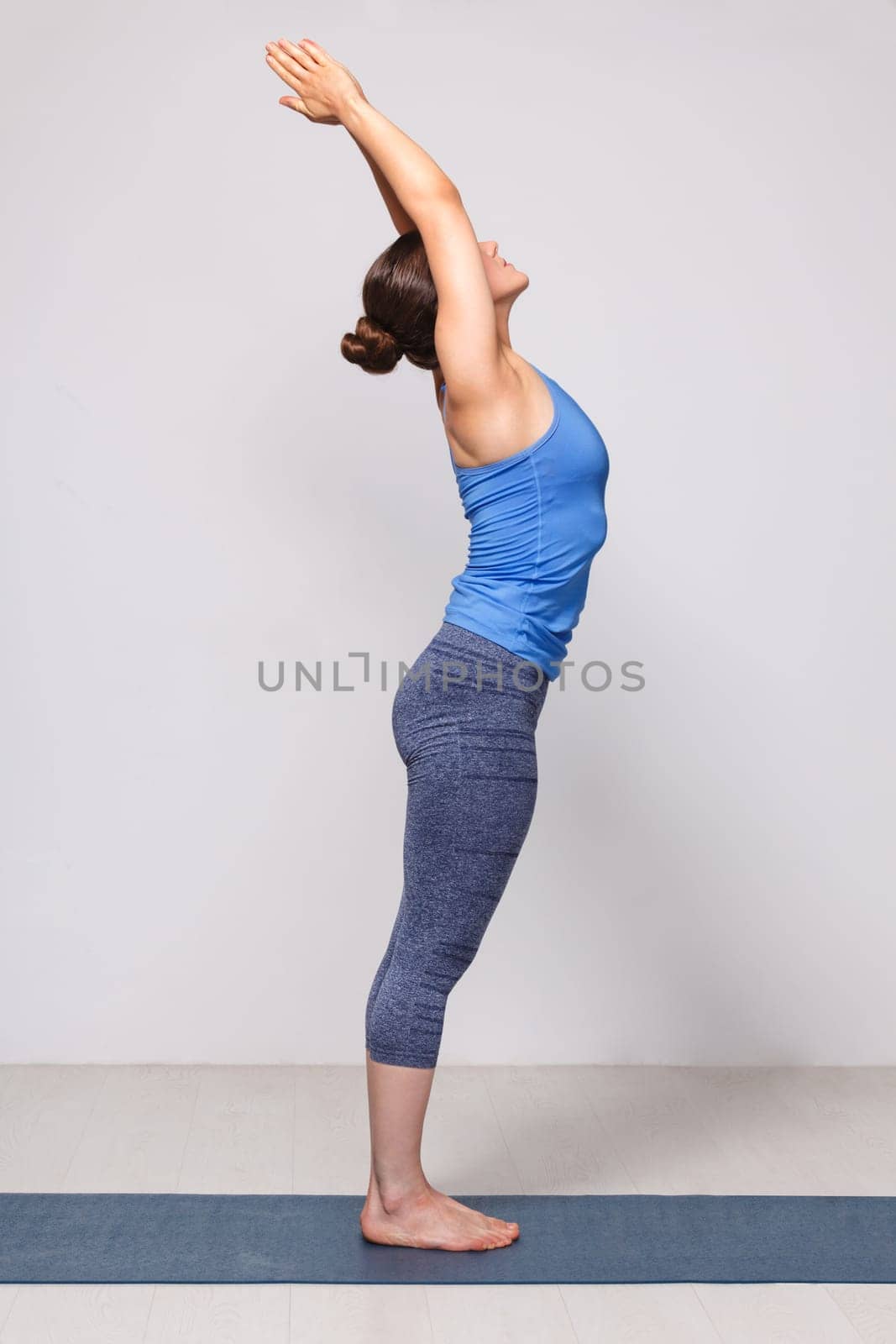Woman doing Hatha Yoga asana Tadasana - Mountain pose with stretched hands on yoga mat in studio on grey bagckground