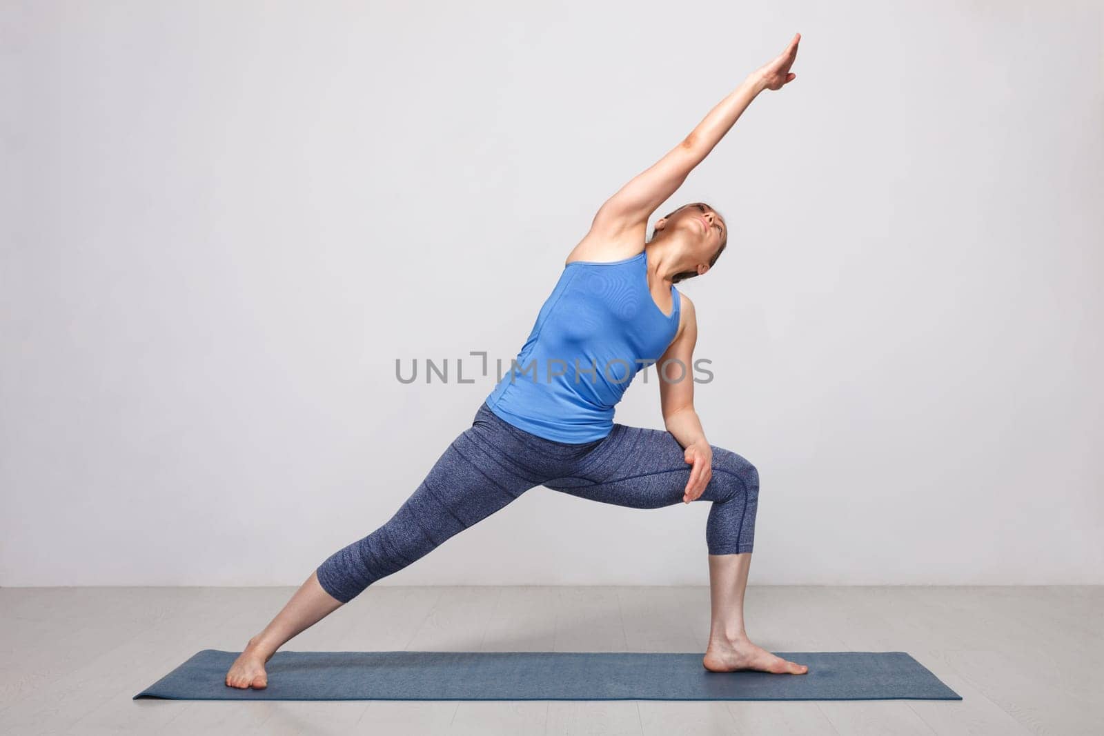 Woman doing Ashtanga Vinyasa yoga asana Utthita parsvakonasana - extended side angle pose beginner variation on yoga mat on yoga mat in studio on grey bagckground