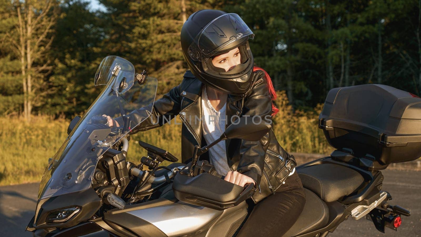 Young biker girl riding touring motorbike on forest road. Beautiful woman with red hair in leather jacket sitting on motorcycle in the park