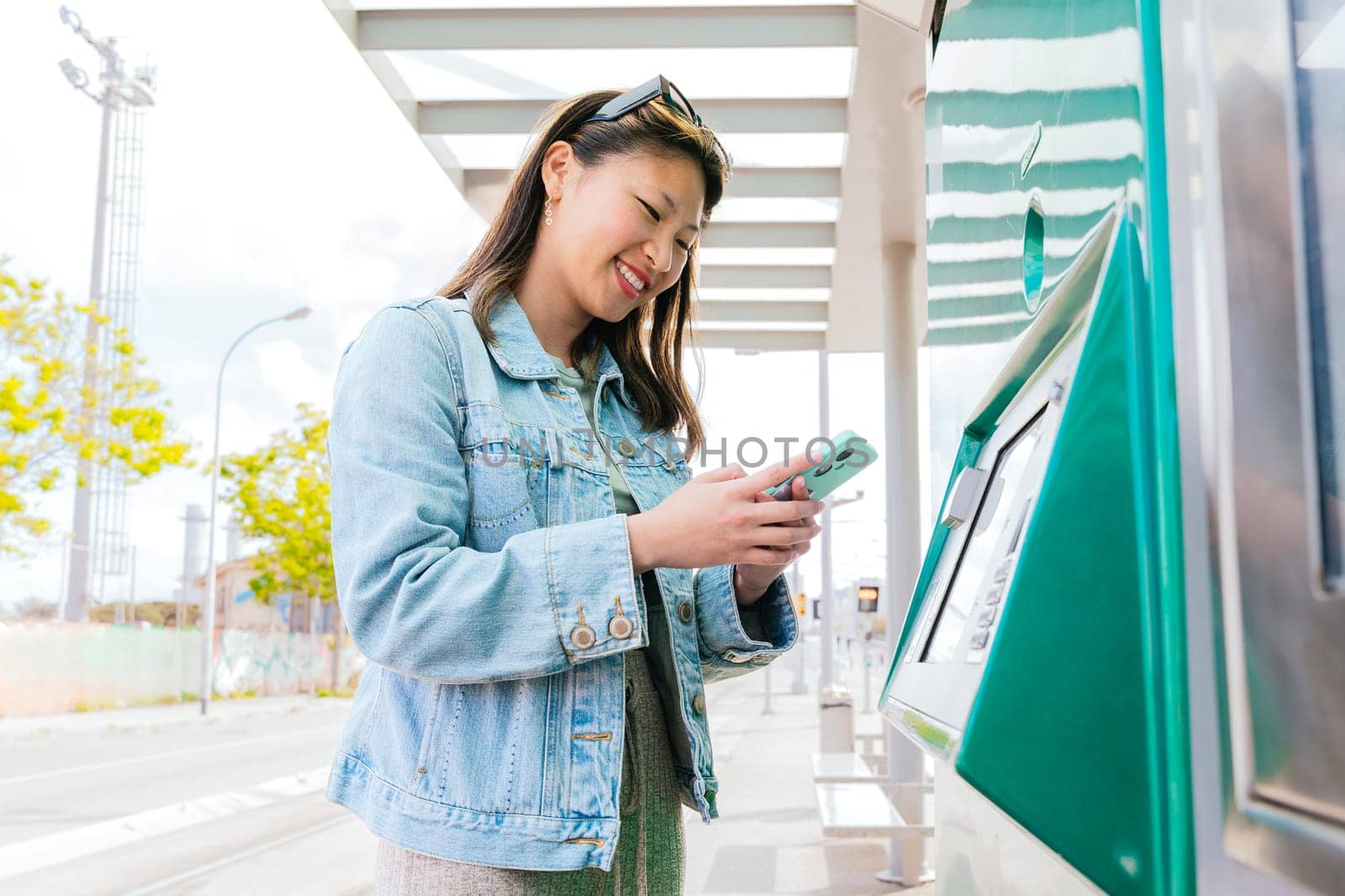Cheerful young woman in a denim jacket using a ticket machine at a tram stop while holding her smartphone. She interacts with the touchscreen in an urban setting, preparing for her journey.