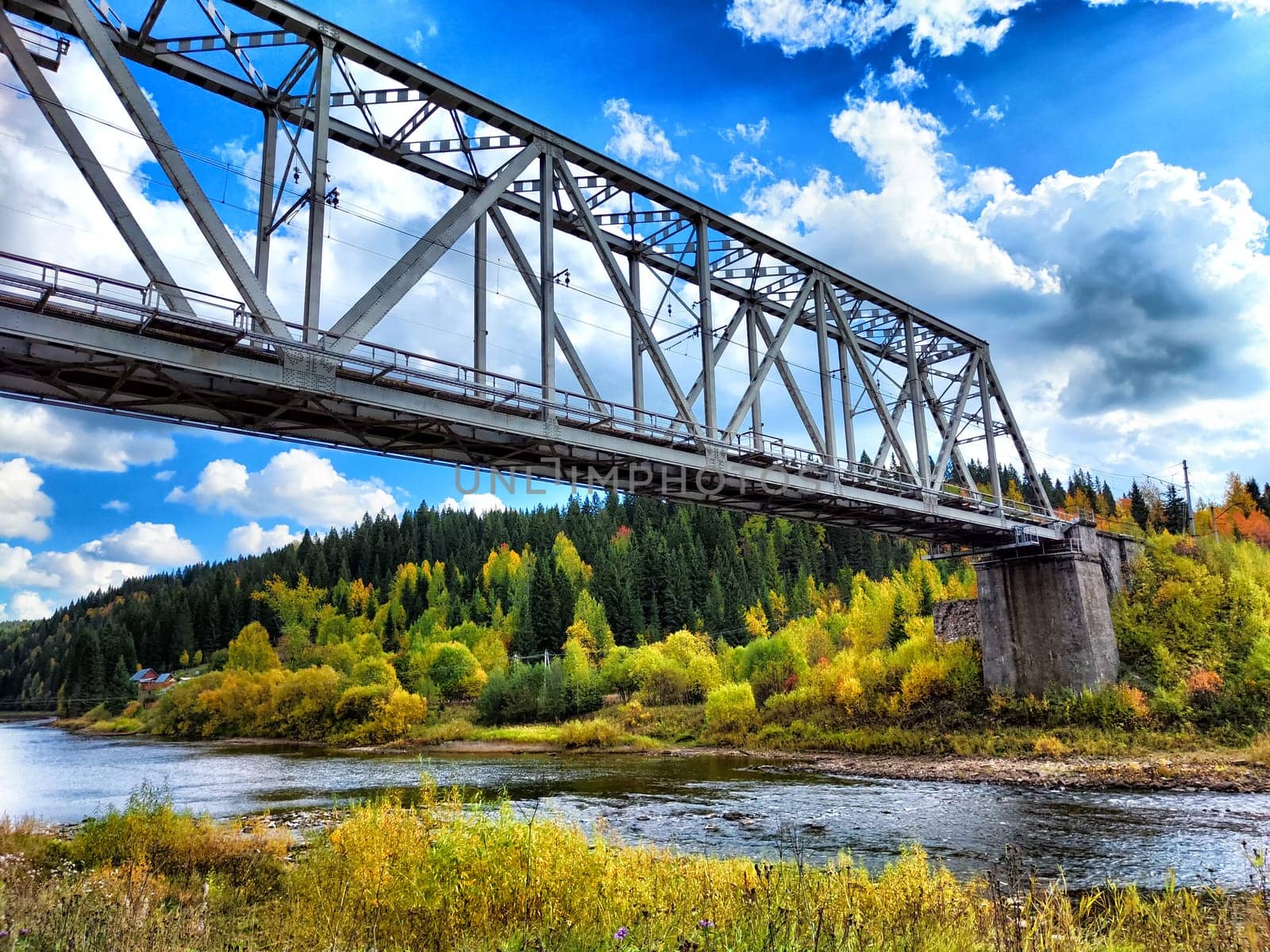 A sturdy metal bridge spans a flowing river, framed by vibrant autumn trees and a clear blue sky filled with clouds.
