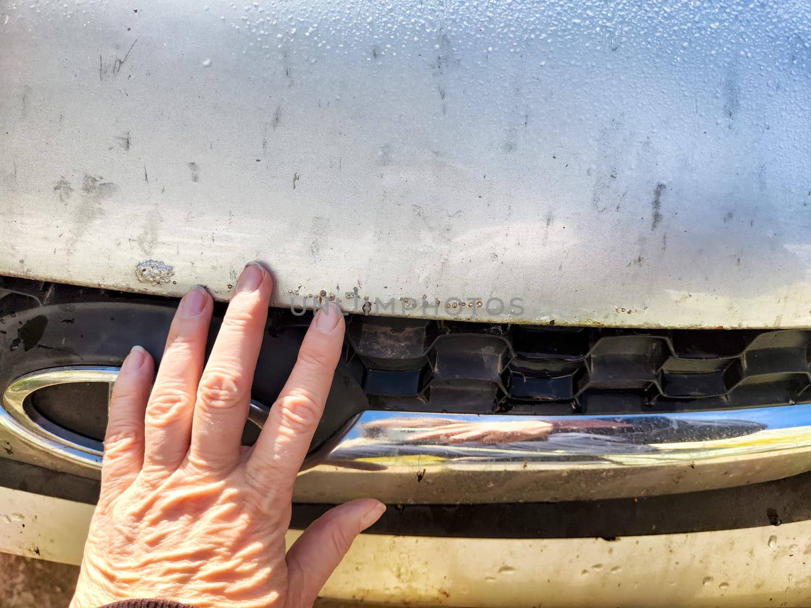 A person examines rust and scratches on silver car parked in a bright outdoor setting, focusing on the damaged area