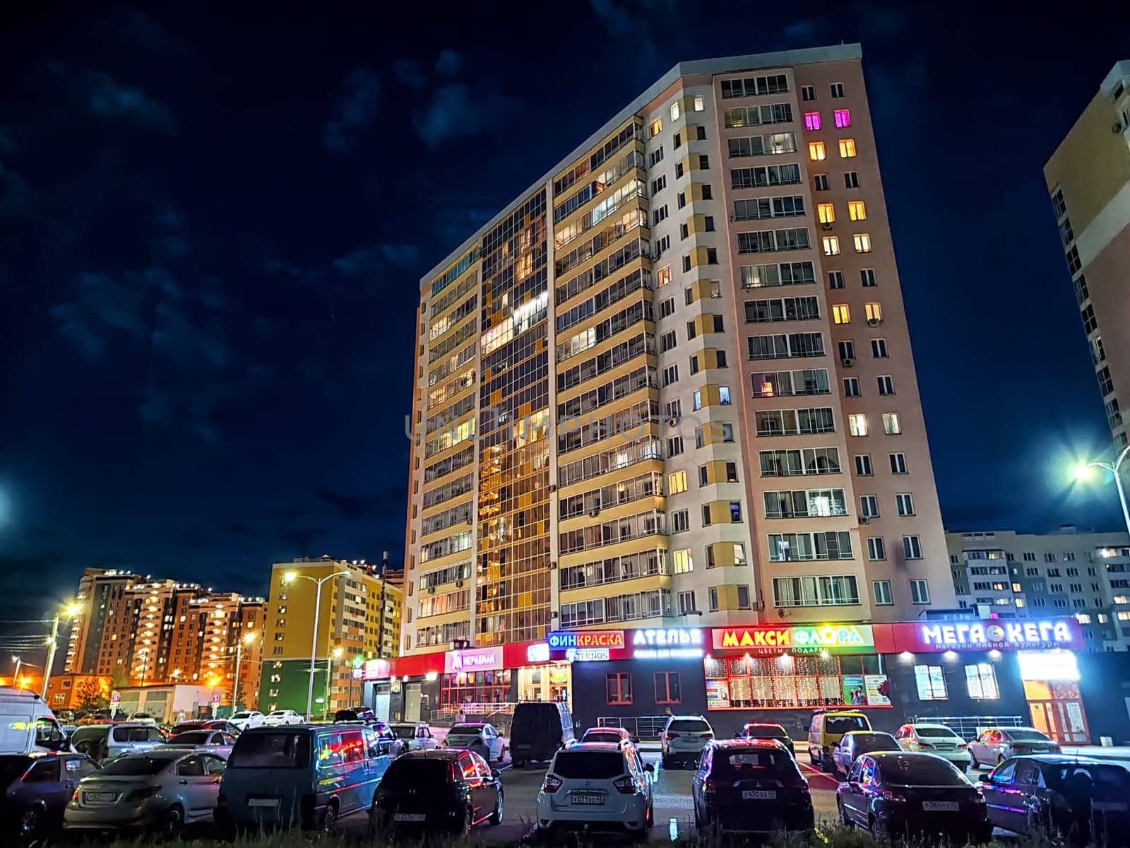 Kirov, Russia - August 11, 2023 - Apartment buildings glow at night with lights in the windows, surrounded by parked cars in a lively urban environment.