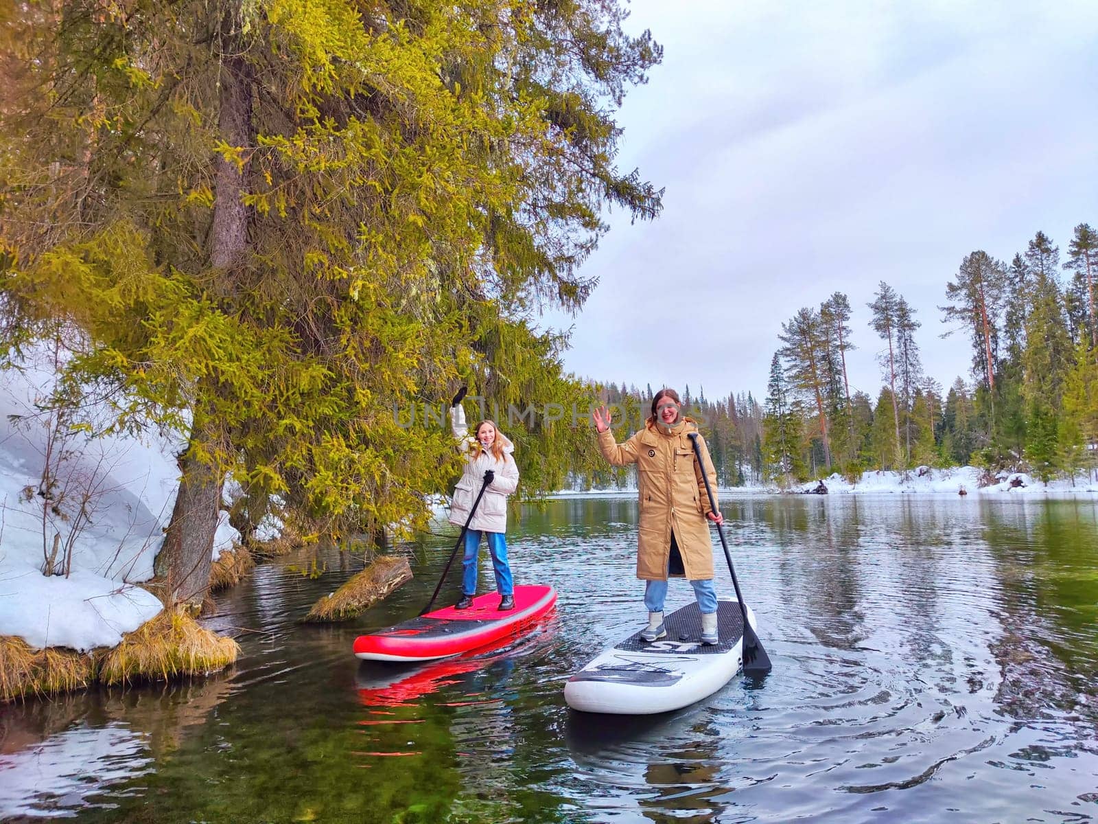 Kirov, Russia - March 14, 2024: Friends enjoy paddle boarding on a calm river, surrounded by snow-covered trees and a peaceful landscape in early spring.
