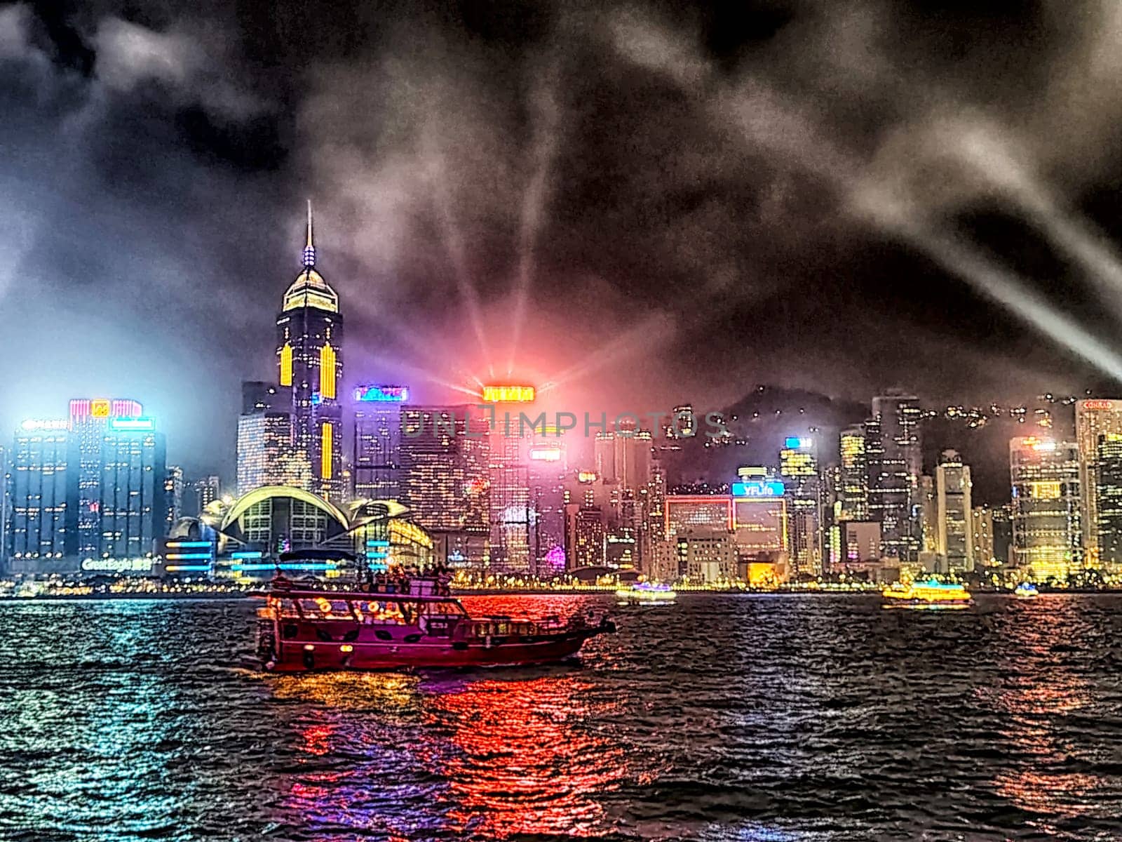 Hong Kong - April 05, 2024: A ship glides through Victoria Bay, illuminated by the colorful lights of Hong Kong's skyscrapers. The skyline sparkles as beams of light dance across the night sky, creating a dynamic atmosphere.