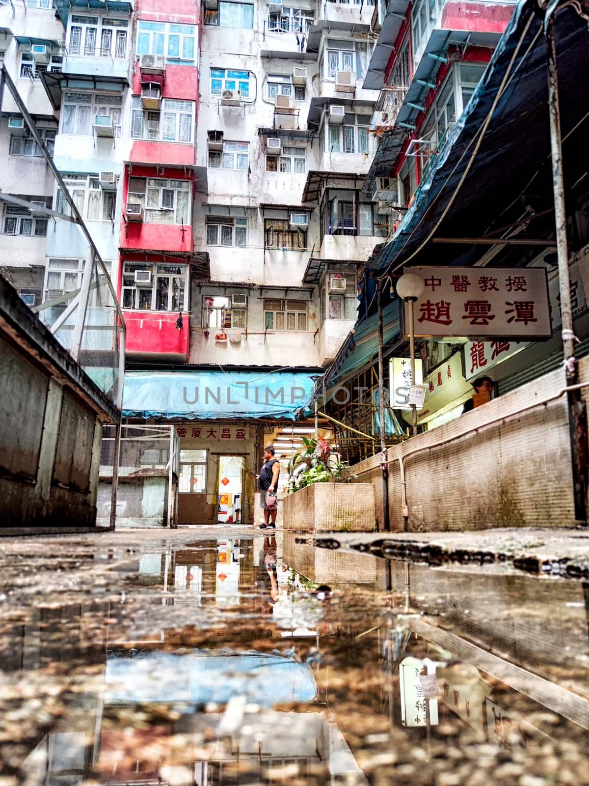 Hong Kong, China - April 06, 2024 - Tourists walk through a narrow street while admiring the towering monster apartment building surrounded by reflections in puddles. The monster House.