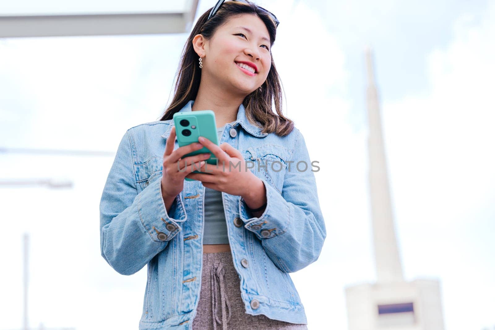 Cheerful young woman in a denim jacket holding her smartphone while looking up with a happy expression. Modern urban setting, representing digital connectivity and city lifestyle.