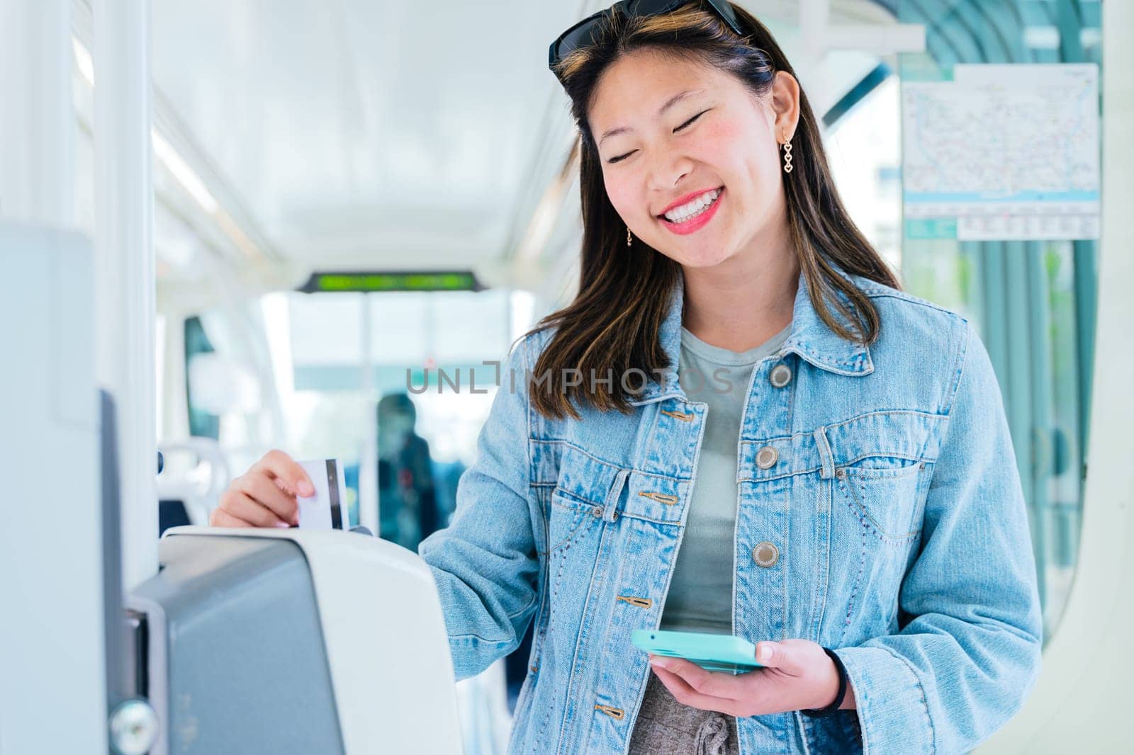 Cheerful young woman in a denim jacket validating her ticket inside public transport while holding a smartphone. She smiles as she uses the modern electronic system in an urban commuting setting.