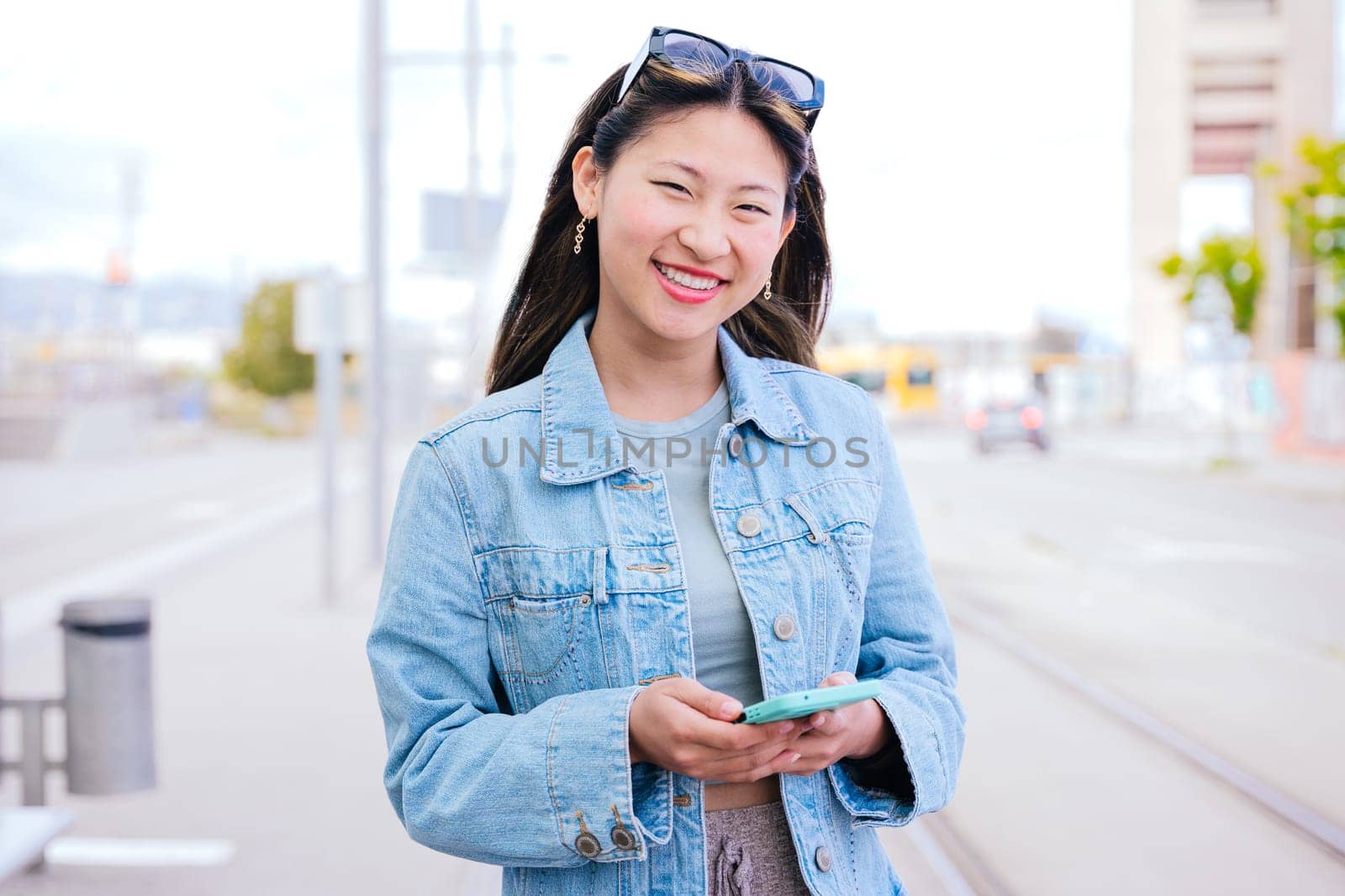 Cheerful young woman in a denim jacket holding her smartphone while looking up with a happy expression. Modern urban setting, representing digital connectivity and city lifestyle.