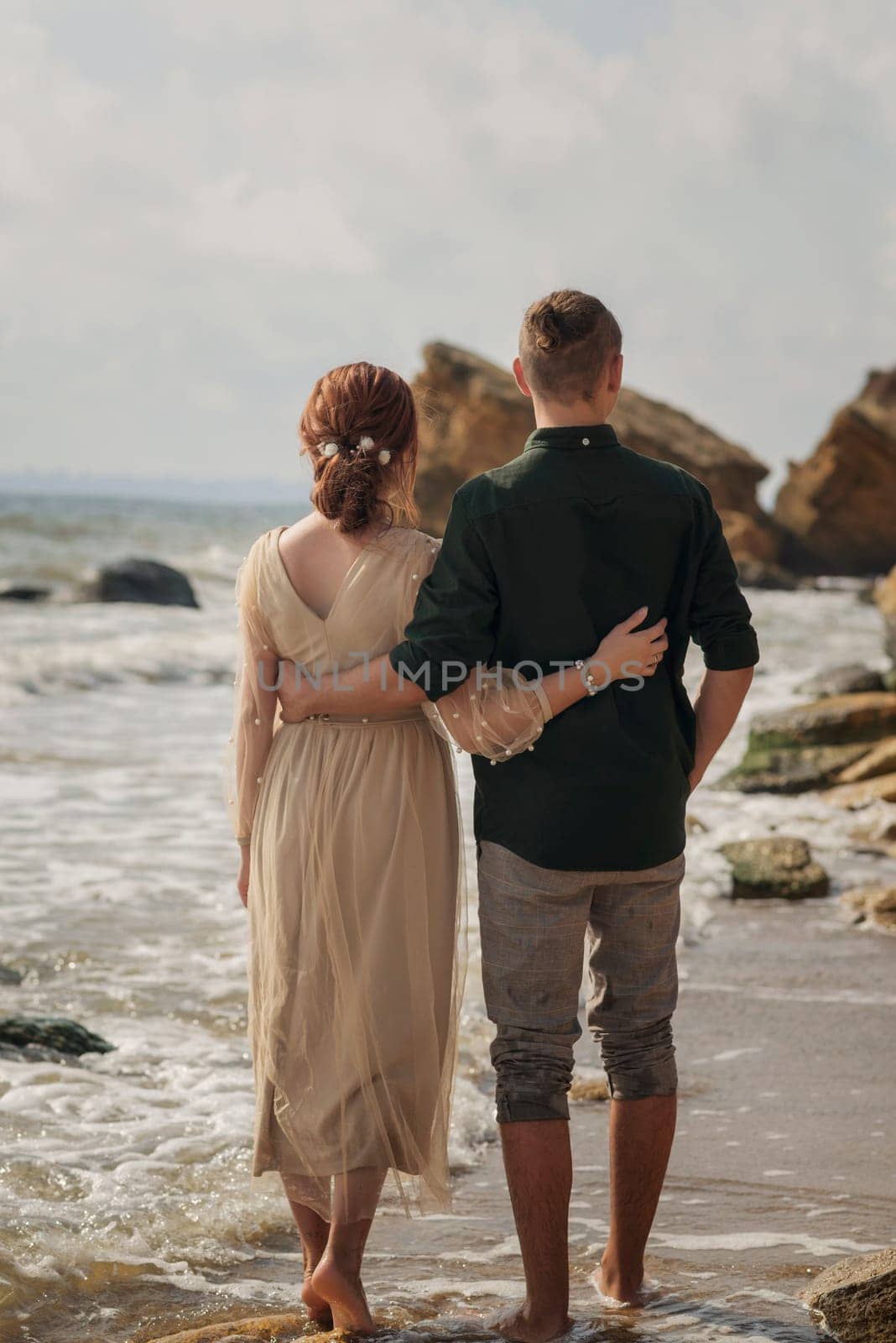 young loving and romantic couple walking along the seashore. view from behind, they hug each other by the waist. and walk along the shore barefoot, rocks in front.