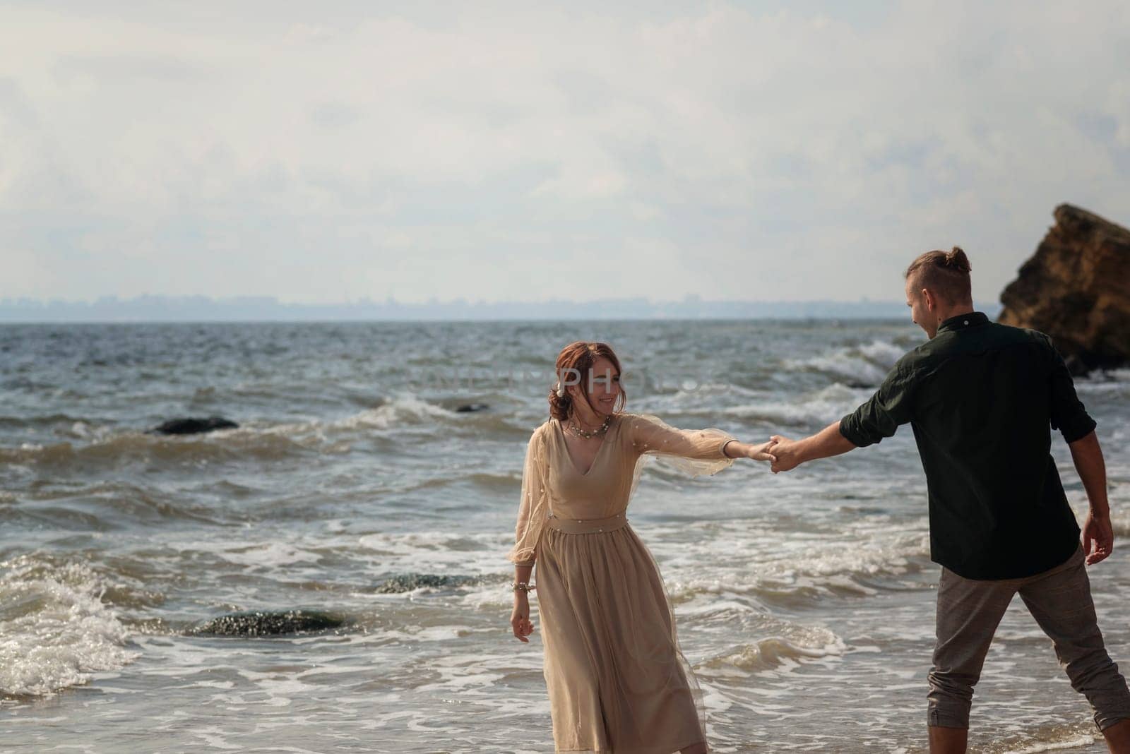 lovers and romantic newlyweds having fun on the seashore. couple dancing on the seashore. they hold hands and look at each other. girl wearing beige dress, wind blowing their hair