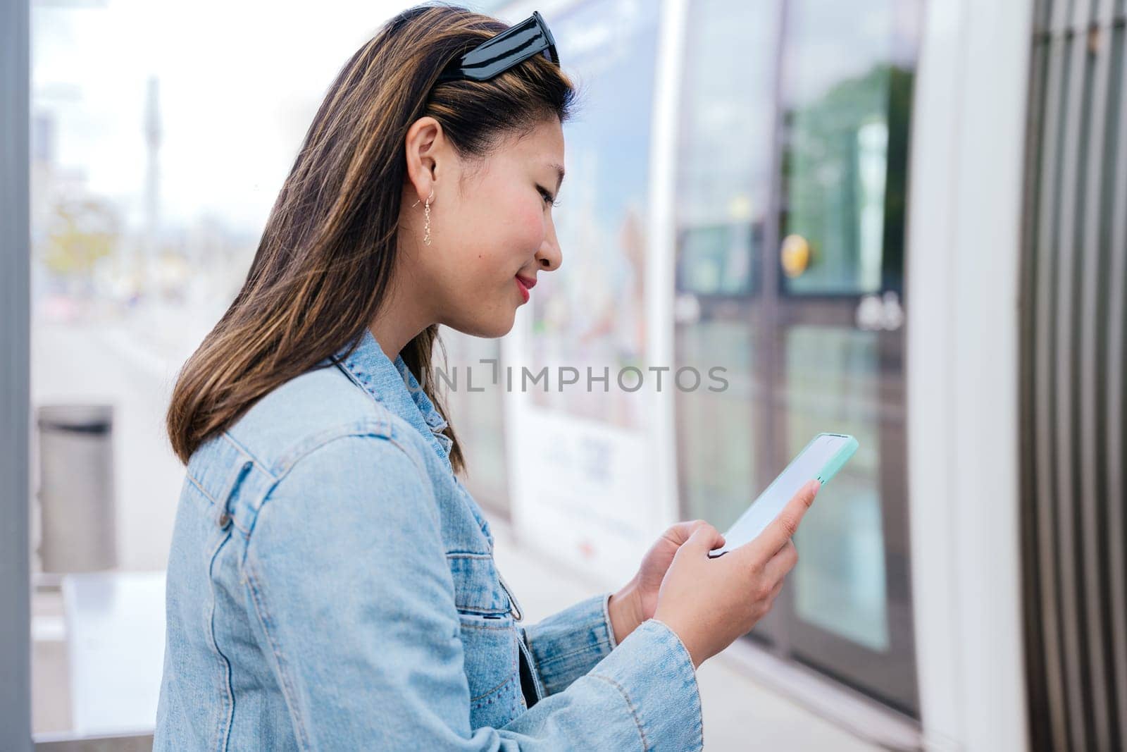 Side view of a young woman in a denim jacket using her smartphone while waiting to board a tram. Modern urban transportation and digital connectivity concept.