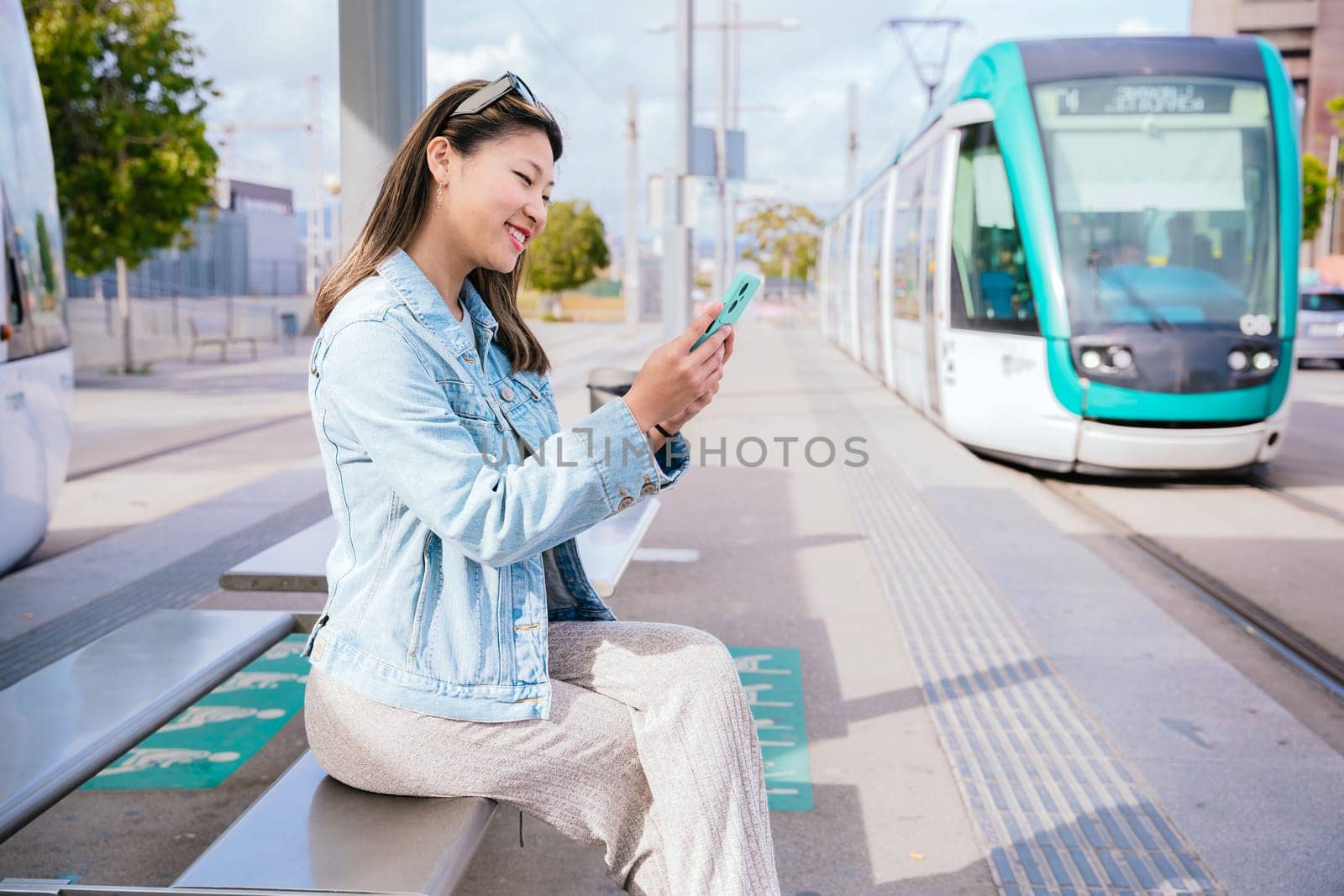 Cheerful young woman in a denim jacket sitting at a tram stop, using her smartphone while waiting for the approaching tram. Modern urban transportation and digital lifestyle concept.