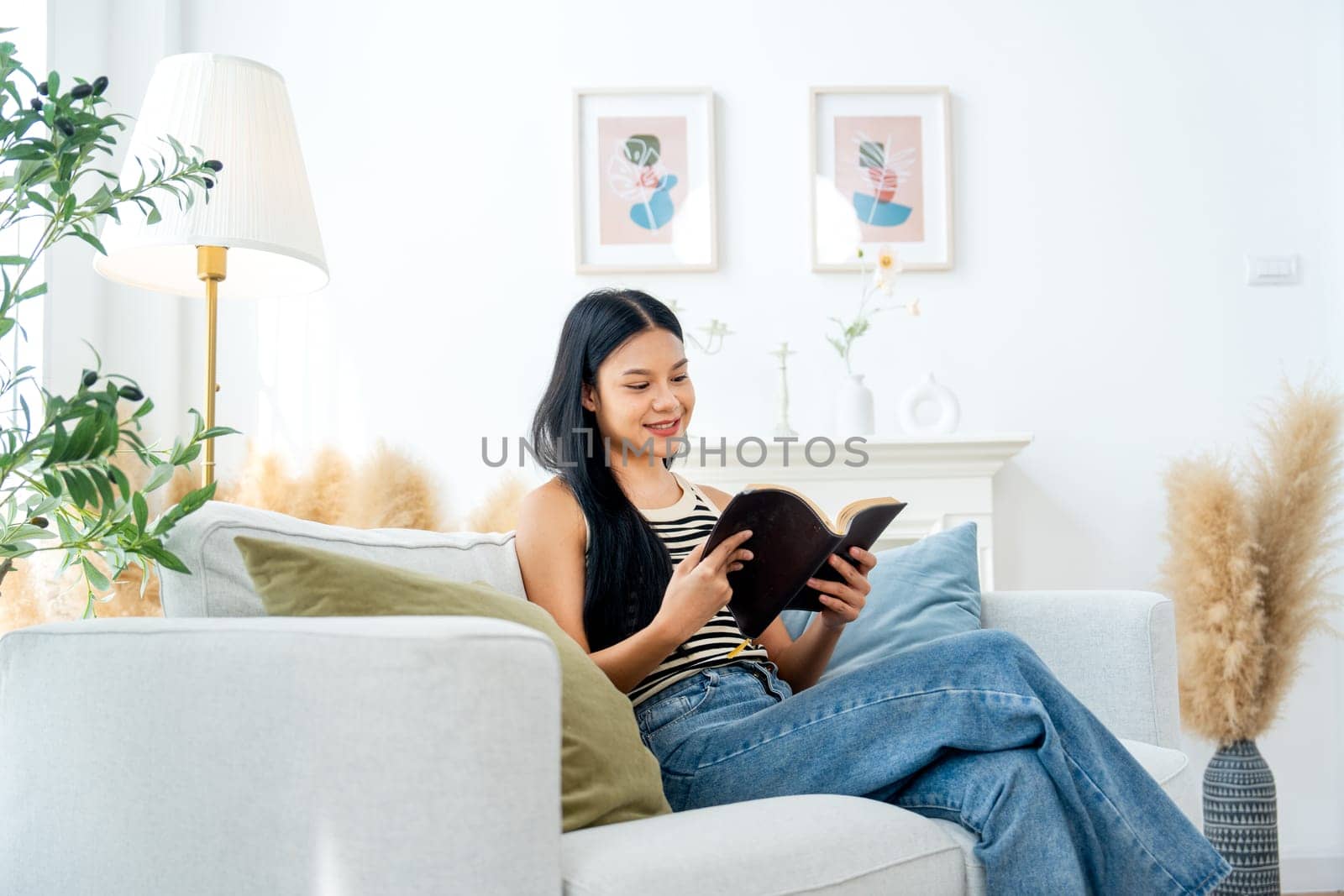 Beautiful Asian woman sit on sofa and enjoy to read the book with relax and happiness in living room with day light.