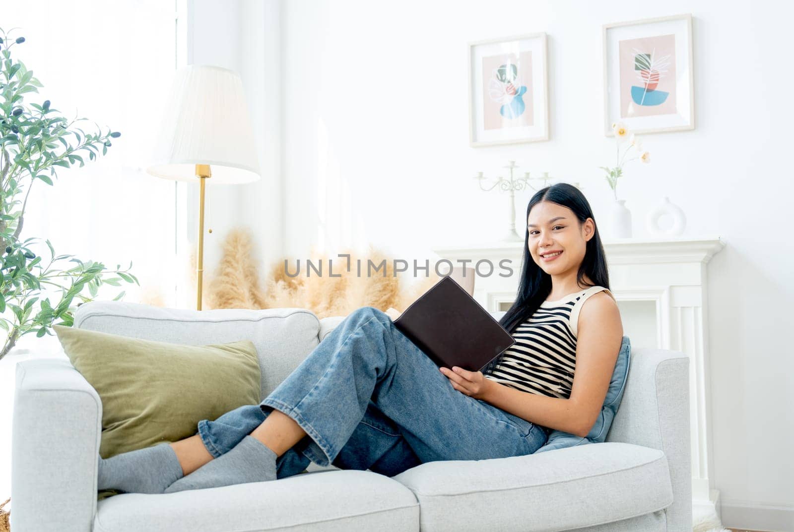Side view and wide shot of beautiful Asian woman lean on sofa also enjoy to read the book and lood at camera with happiness on day light.