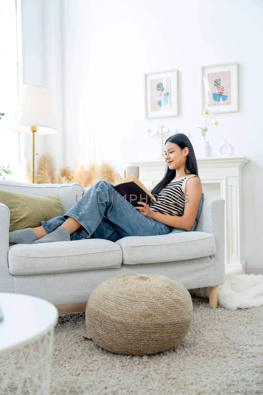 Vertical image of beautiful Asian woman lean on sofa hold a cup of tea also enjoy to read the book with happiness on day light.