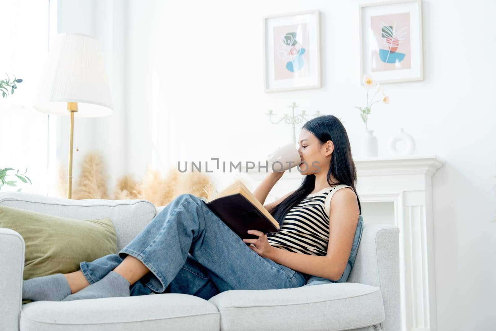 Beautiful Asian woman lean on sofa drink tea also enjoy to read the book with happiness in living room with day light.