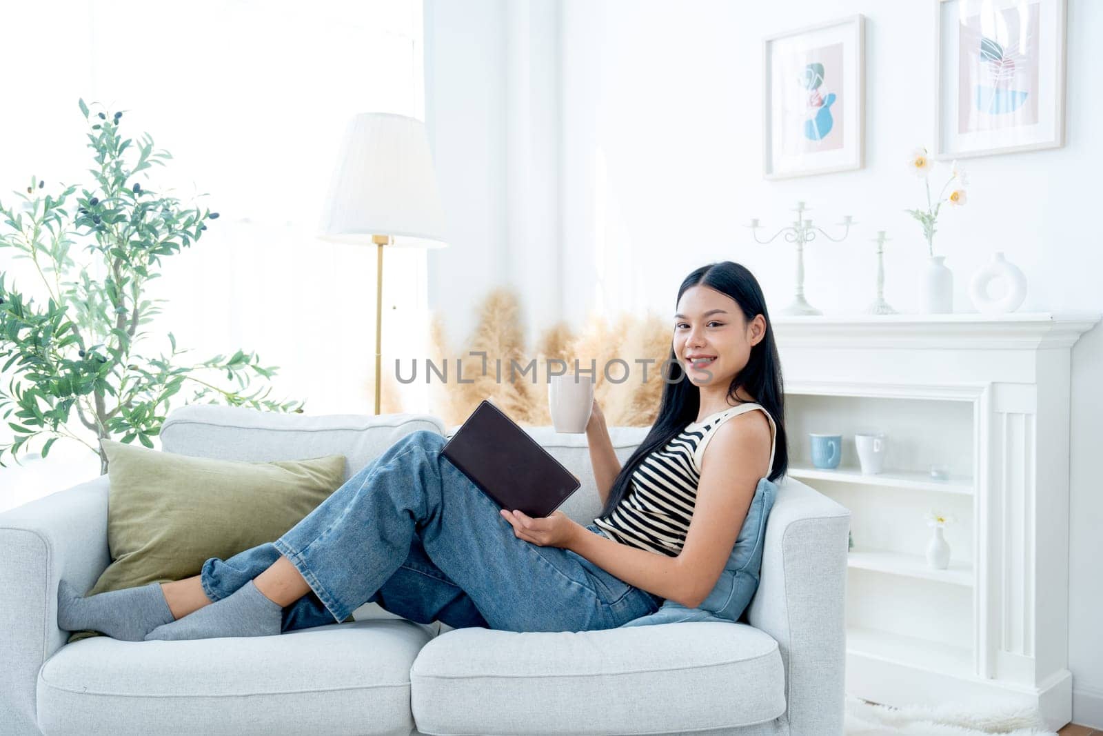 Side view and wide shot of beautiful Asian woman lean on sofa hold a cup of tea also enjoy to read the book and lood at camera with happiness on day light.