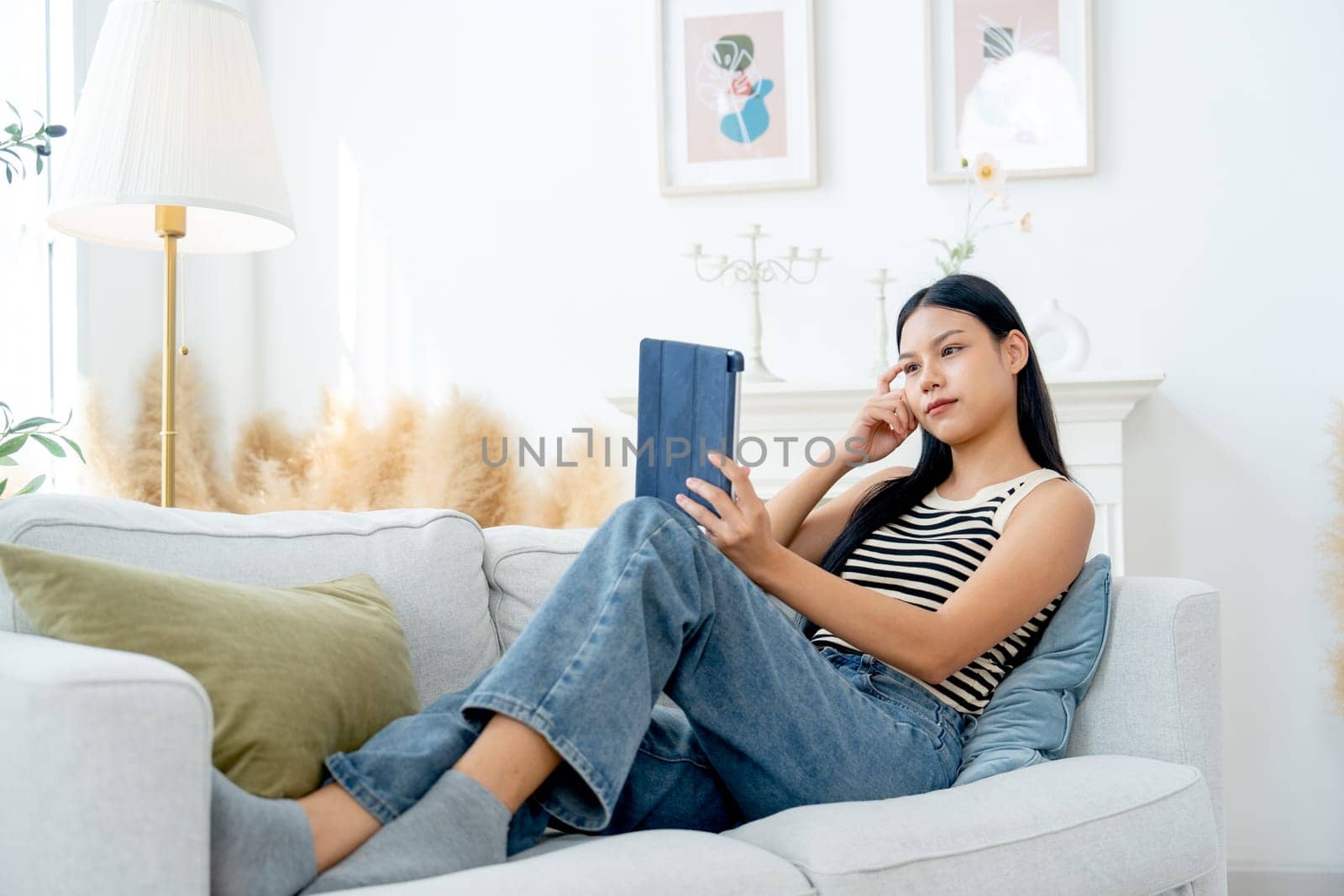 Wide shot of beautiful Asian woman lean on sofa also enjoy to use tablet in living room and express thinking action with day light.