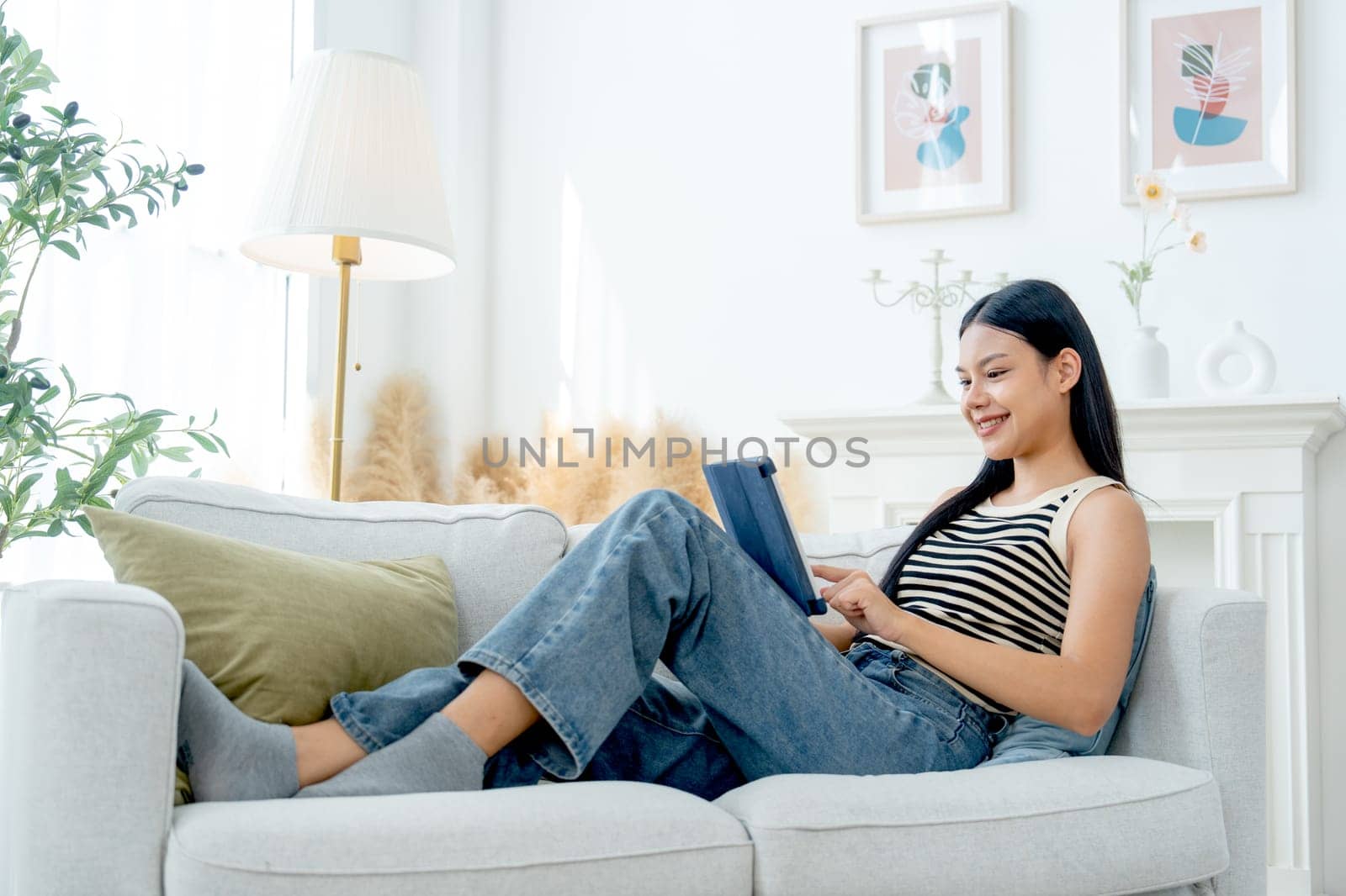 Side and high view beautiful Asian woman lean on sofa also enjoy to use tablet in living room with day light.