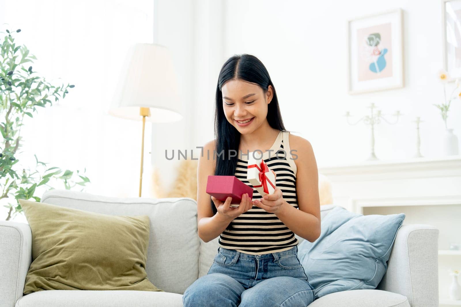 Beautiful Asian woman sit on sofa in living room and hold box of present also look at the box and express happiness with  day light.