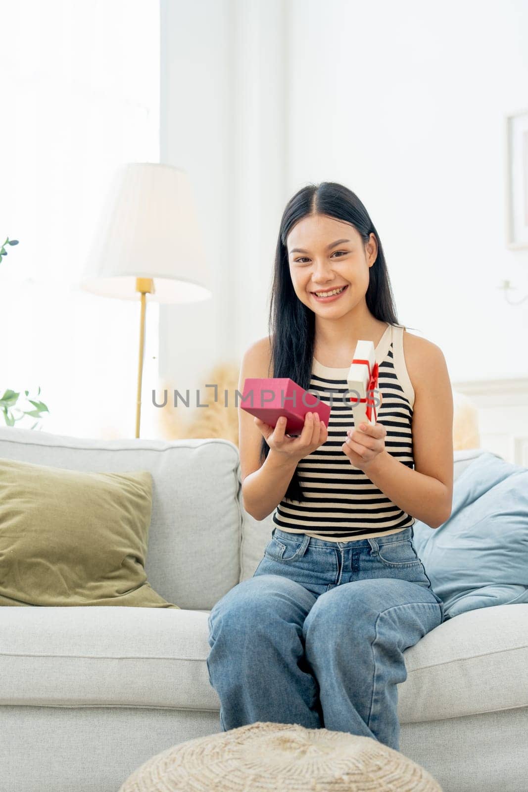 Vertical image of beautiful Asian woman sit on sofa in living room and hold box of present also look at camera with smiling and day light.