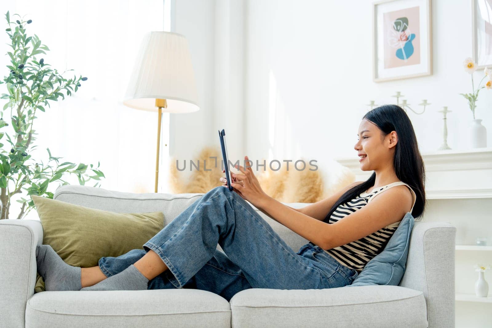 Side view and wide shot beautiful Asian woman lean on sofa also enjoy to use tablet in living room with day light.