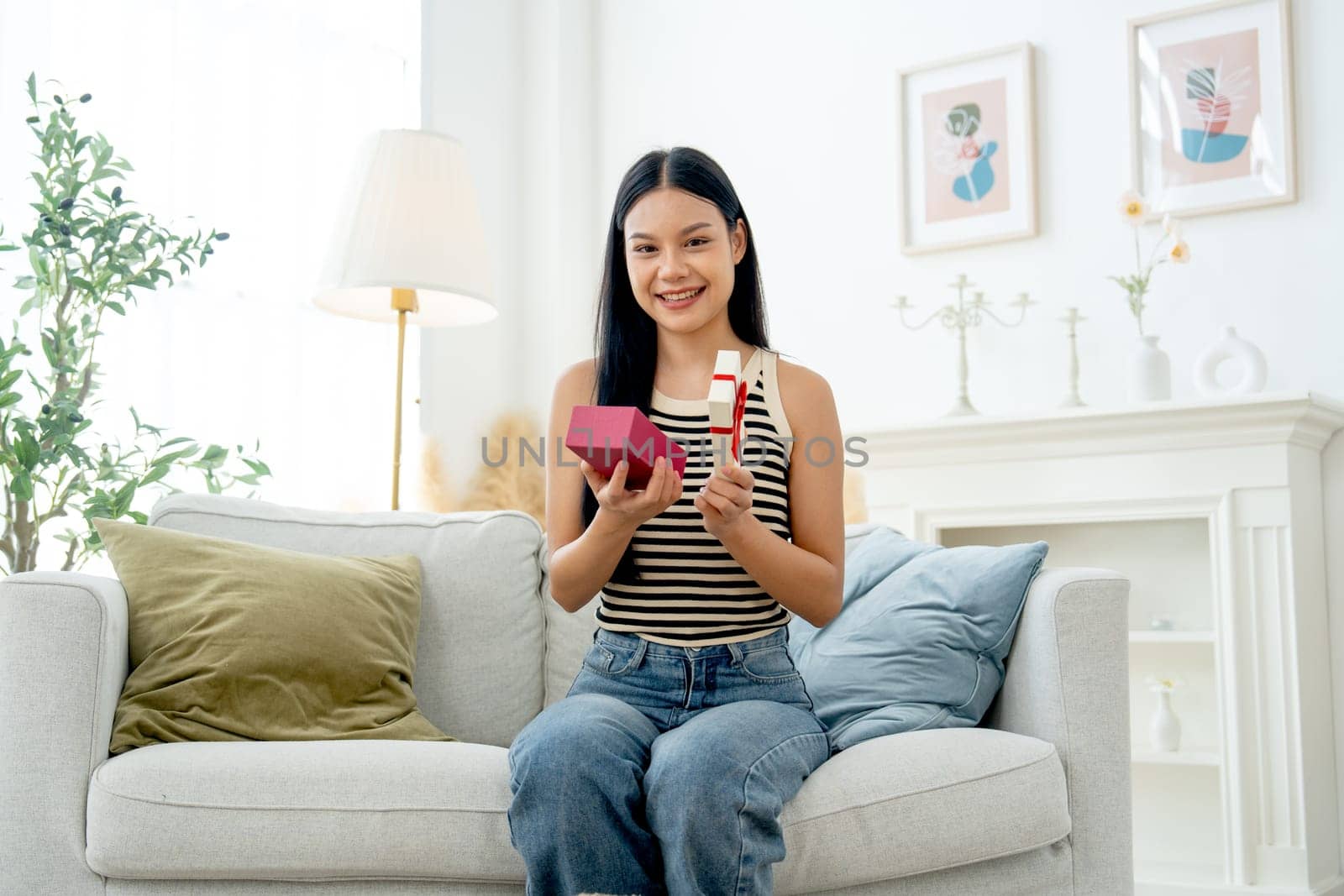 Beautiful Asian woman sit on sofa in living room and hold box of present also look at camera with smiling and day light.