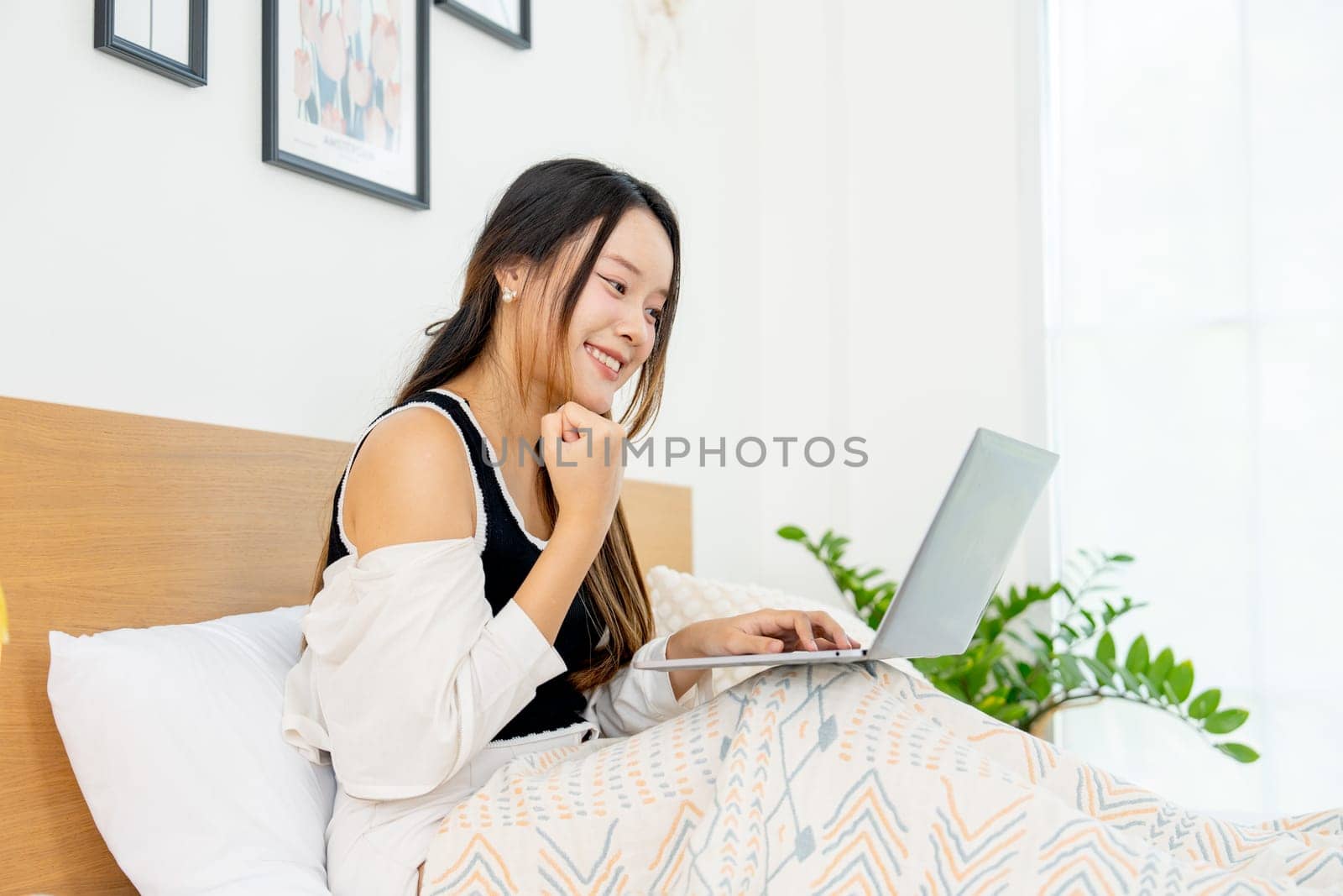 Side view of lovely Asian woman sit on bed and enjoy to use laptop for relaxing in bedroom.