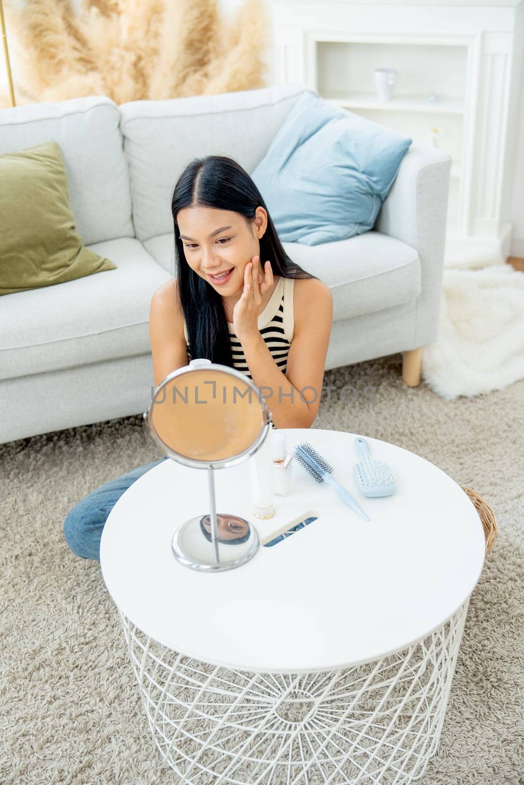 Vertical image and front view beautiful Asian woman sit in front of mirror enjoy to makeup and apply cosmetic on her face with happiness in living room of her house.