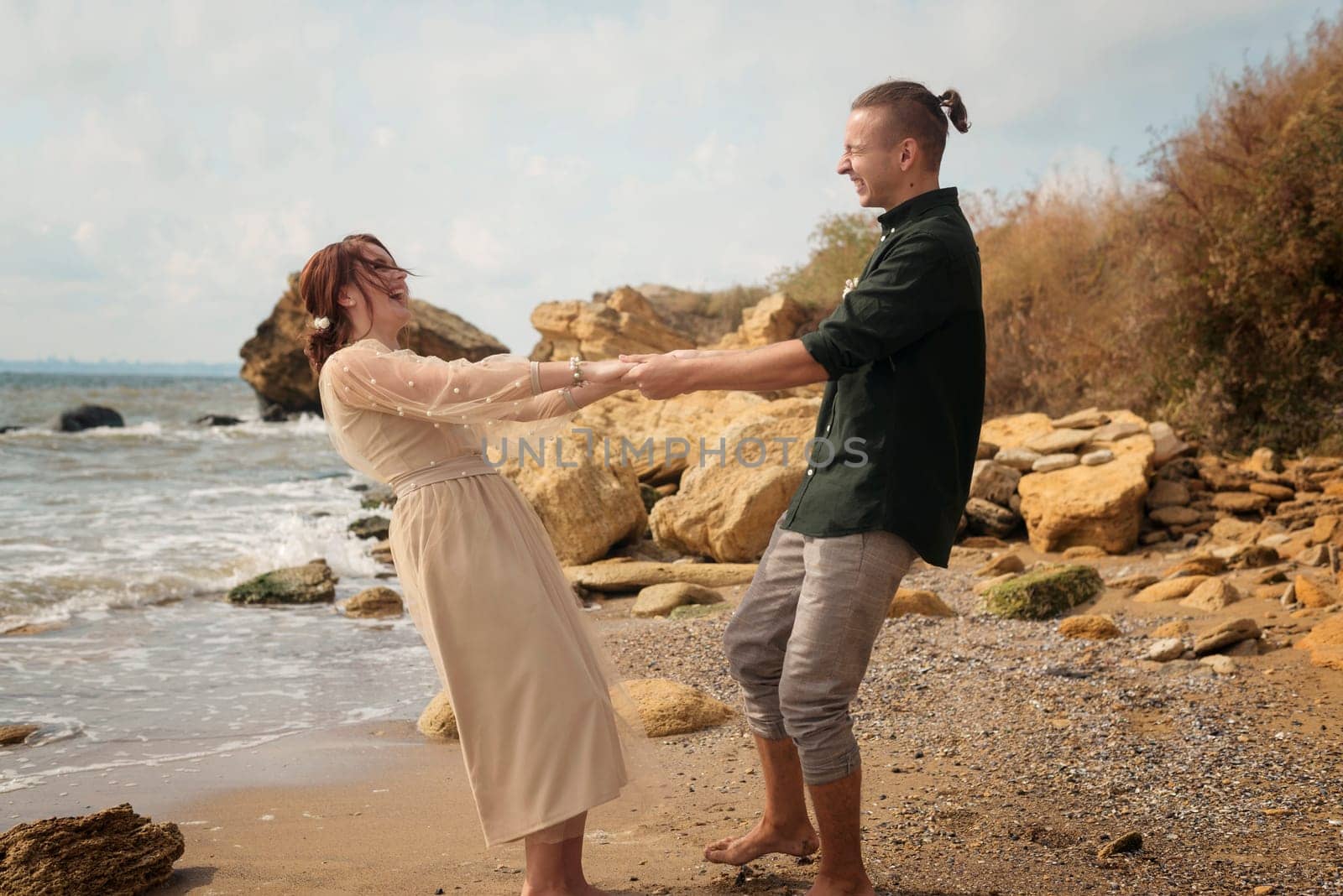 romantic and smiling couple dancing on the seashore. they are spinning barefoot on the seashore. the couple is holding hands, smiling and laughing. the sea and rocks are around them.