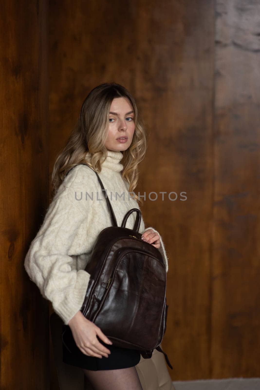 A girl in a warm sweater, a black short skirt, nylon tights, and black shoes is posing with a brown backpack
