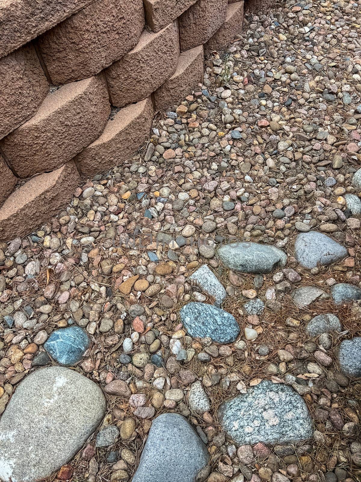 A close-up view of an aging retaining wall made of stacked concrete blocks, surrounded by a bed of decorative stones in various colors and sizes. The landscape shows signs of wear and is ready for renovation to restore its appeal and functionality.