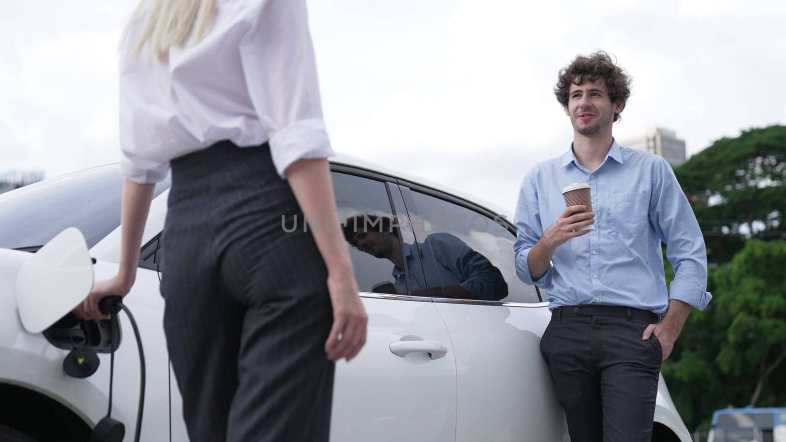 Progressive businessman and businesswoman leaning on EV car and charging point. by biancoblue