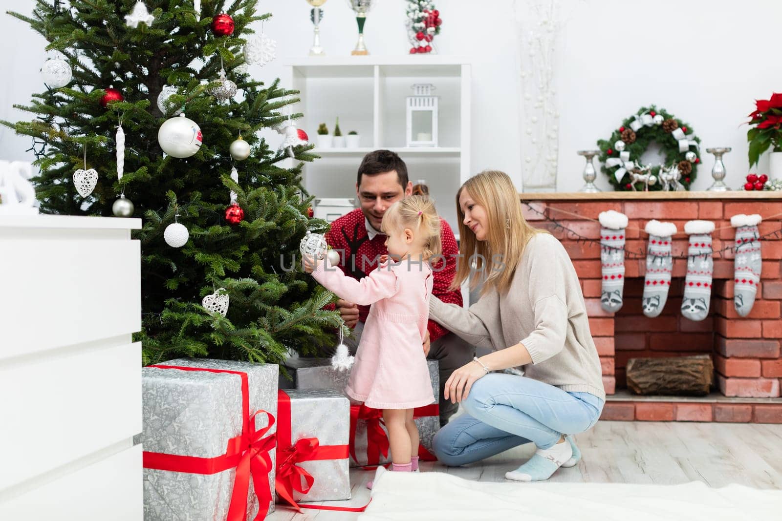 Mom, dad and little daughter are staying together by the Christmas tree. In the background you can see a brick fireplace, on which Christmas socks are hung. Under the Christmas tree lie large boxes of gifts. The boxes are decorated with red ribbon.