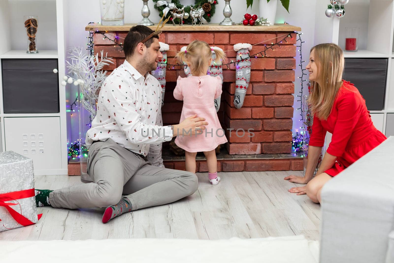 A man and a woman are sitting on the floor by a brick fireplace. Between them stands a little girl. The girl is facing away from the fireplace. The whole family is wearing festive clothes