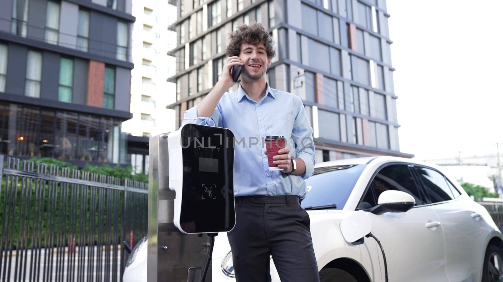 Suit-clad businessman with progressive ambition leaning on his electric vehicle while standing on a charging station with a power cable plug and a renewable energy-powered electric vehicle.