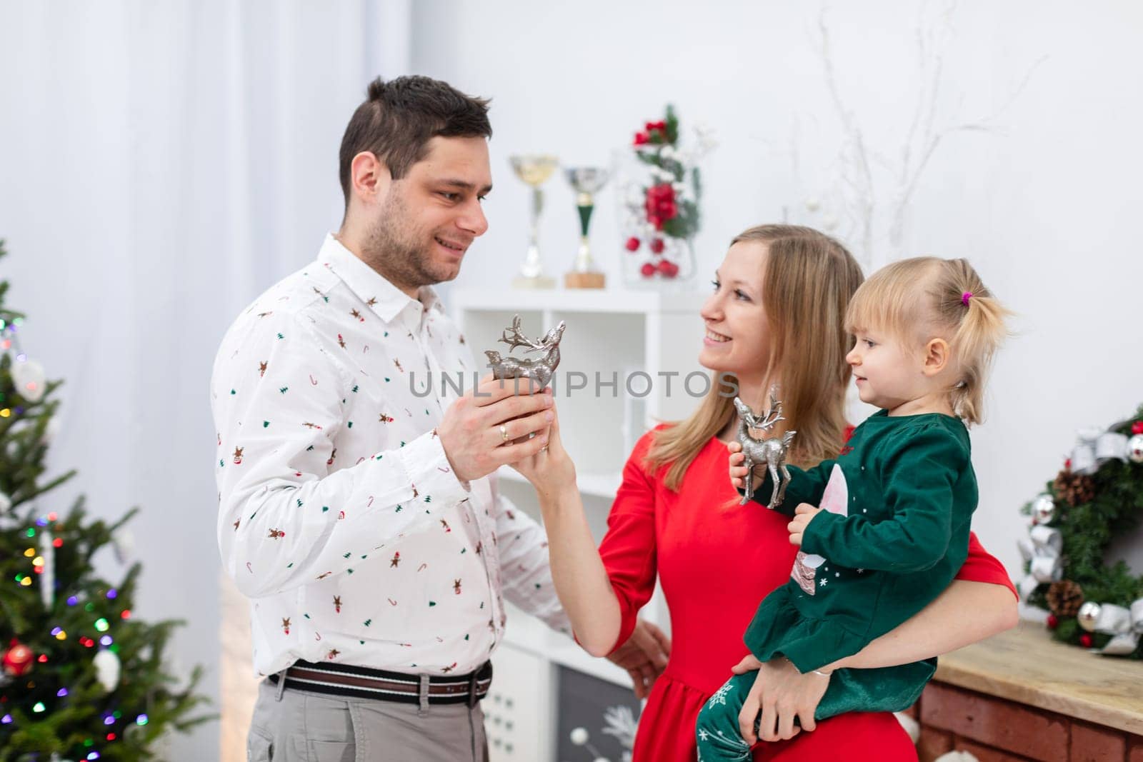 A man, a woman and a little girl stand by a brick fireplace. The fireplace is decorated with Christmas decorations. The woman holds a little girl in her arms. The daughter and father are holding silver Christmas decorations in their hands.