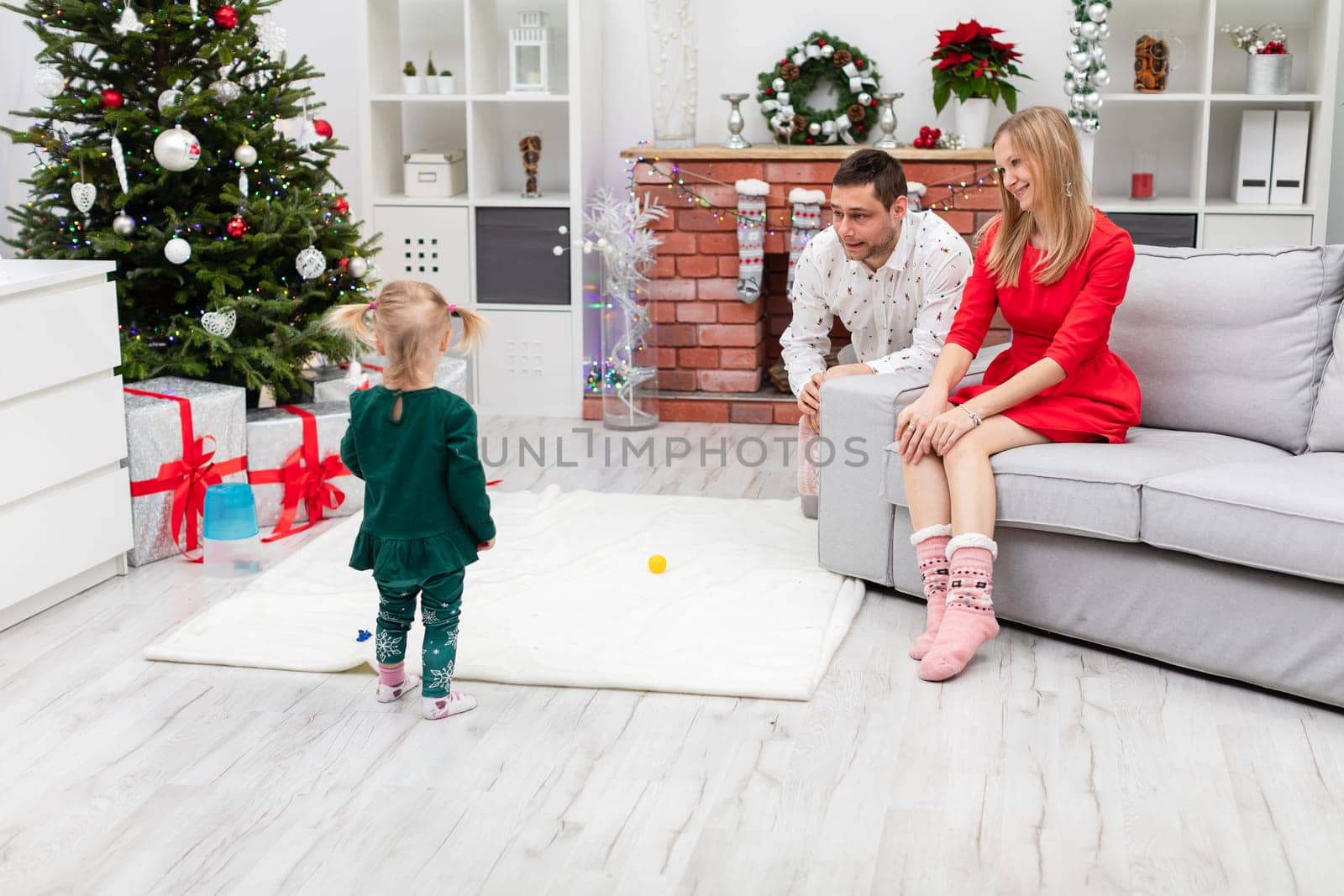 In the foreground a little girl standing with her back. Next to it, a man and a woman are looking at the little girl. The parents and daughter are in a room decorated with Christmas decorations.