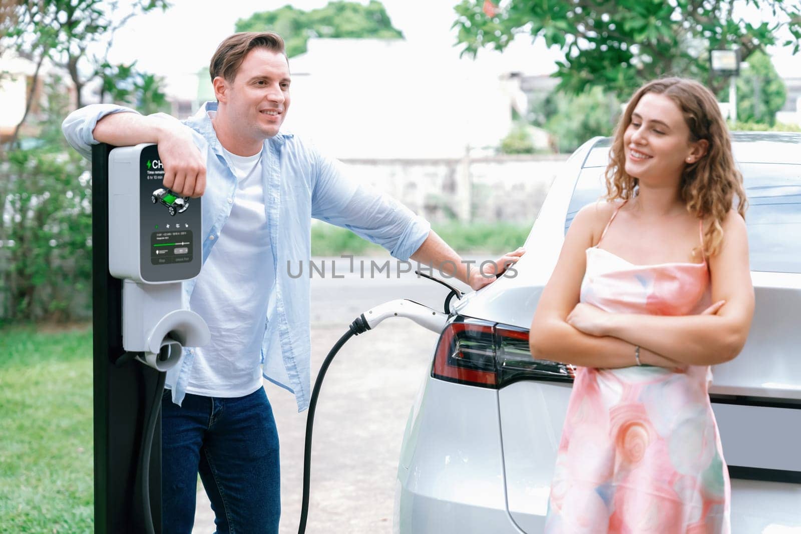 Happy and lovely couple with eco-friendly conscious recharging electric vehicle from EV charging station. EV car technology utilized as alternative transportation for future sustainability. Synchronos