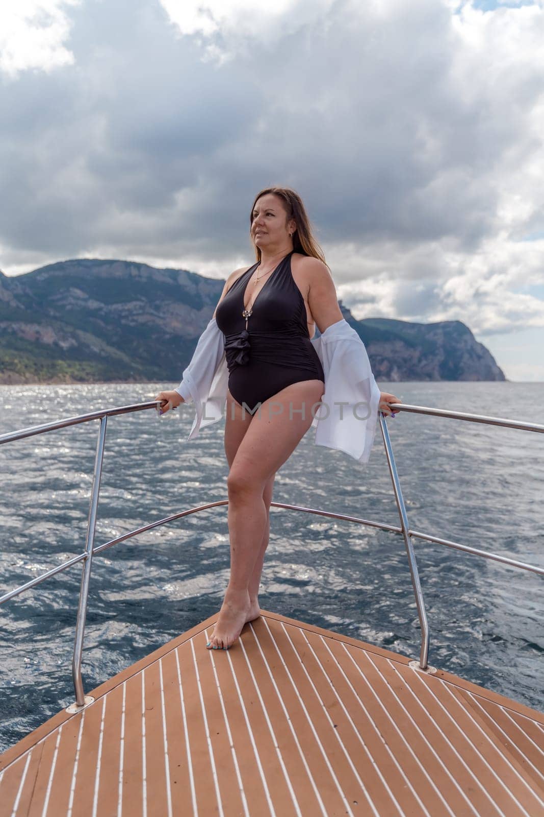 Woman on a yacht. Happy model in a swimsuit posing on a yacht against a blue sky with clouds and mountains by Matiunina