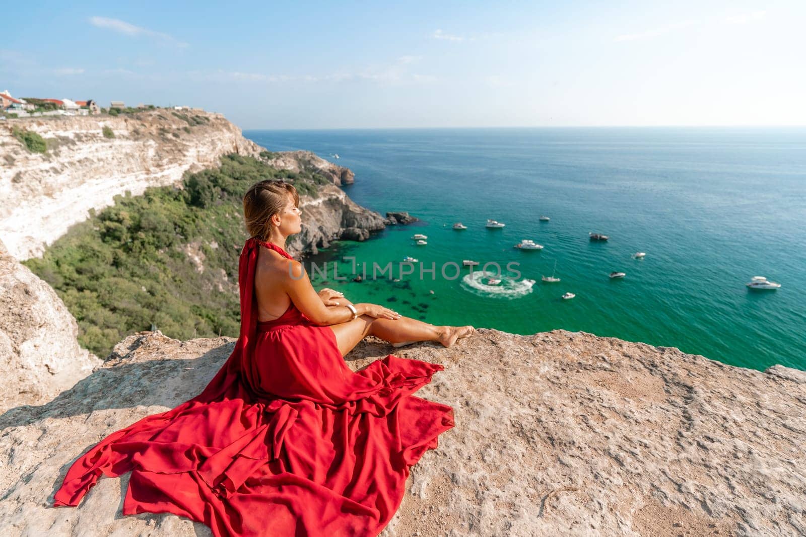 Woman sea red dress yachts. A beautiful woman in a red dress poses on a cliff overlooking the sea on a sunny day. Boats and yachts dot the background