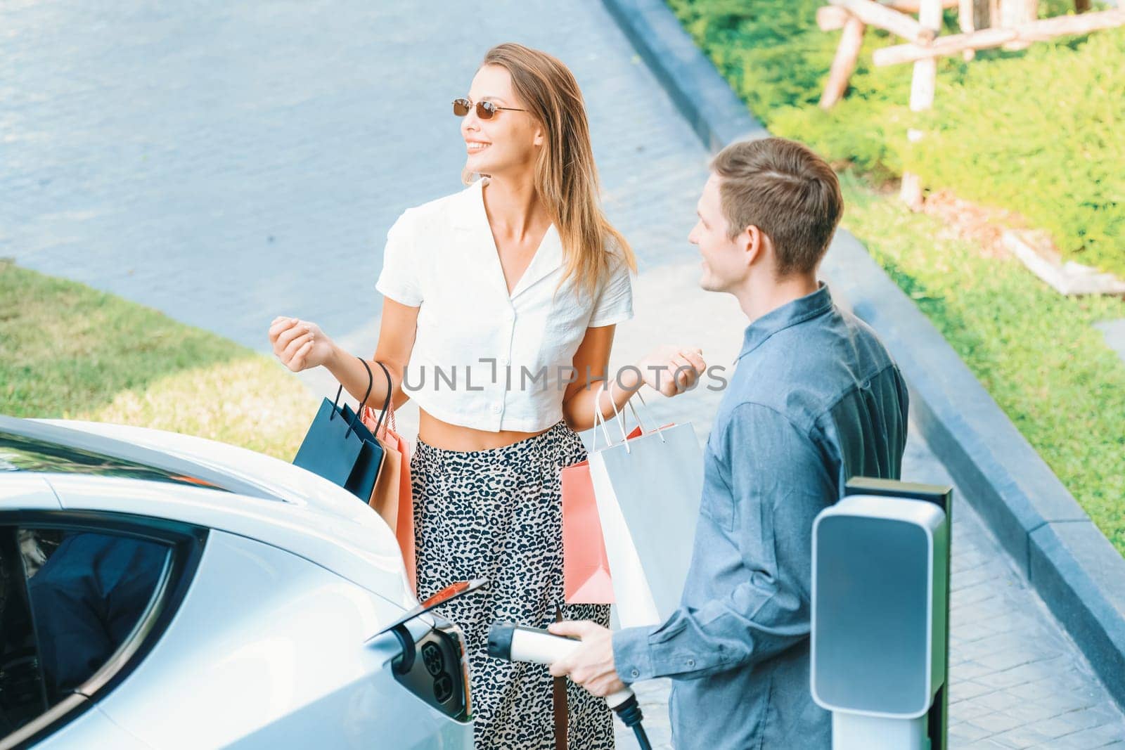 Young couple travel with EV electric car charging in green sustainable city outdoor garden in summer shows urban sustainability lifestyle by green clean rechargeable energy of electric vehicle innards