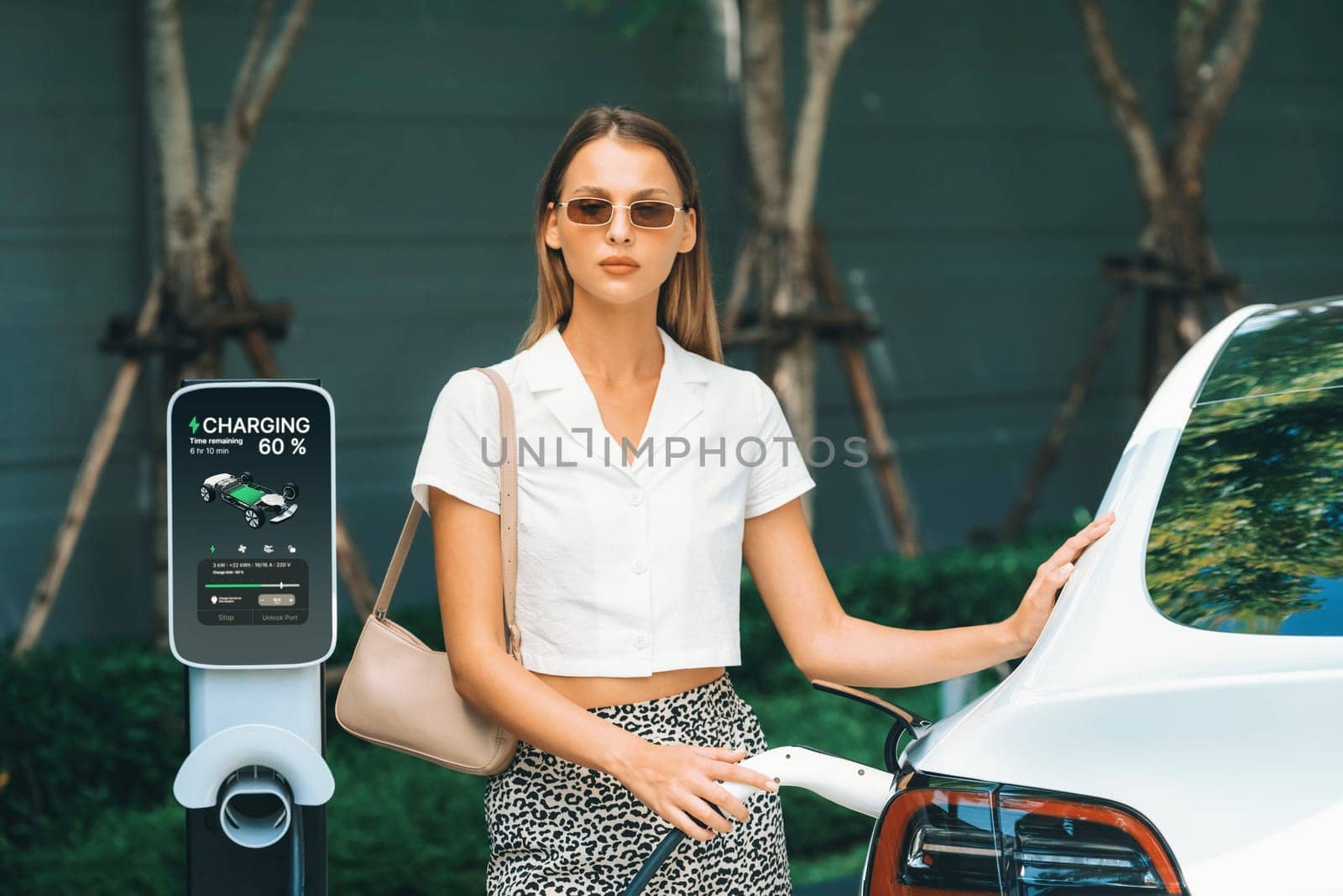 Young woman travel with EV electric car charging in green sustainable city outdoor garden in summer. Urban sustainability lifestyle by green clean rechargeable energy of electric BEV vehicle innards