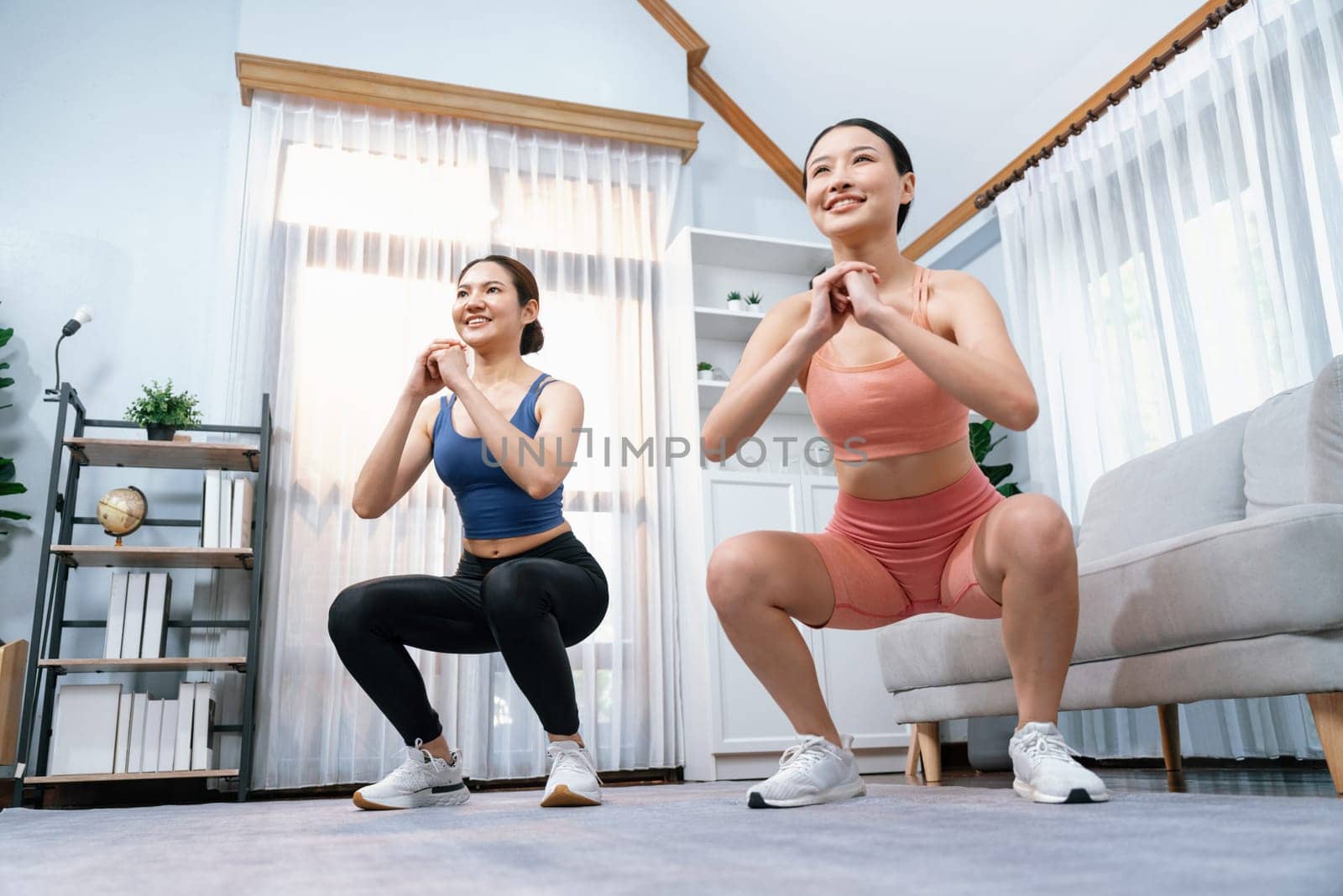 Vigorous energetic woman with trainer or workout buddy doing exercise at home. Young athletic asian woman strength and endurance training session as home workout routine with squat.