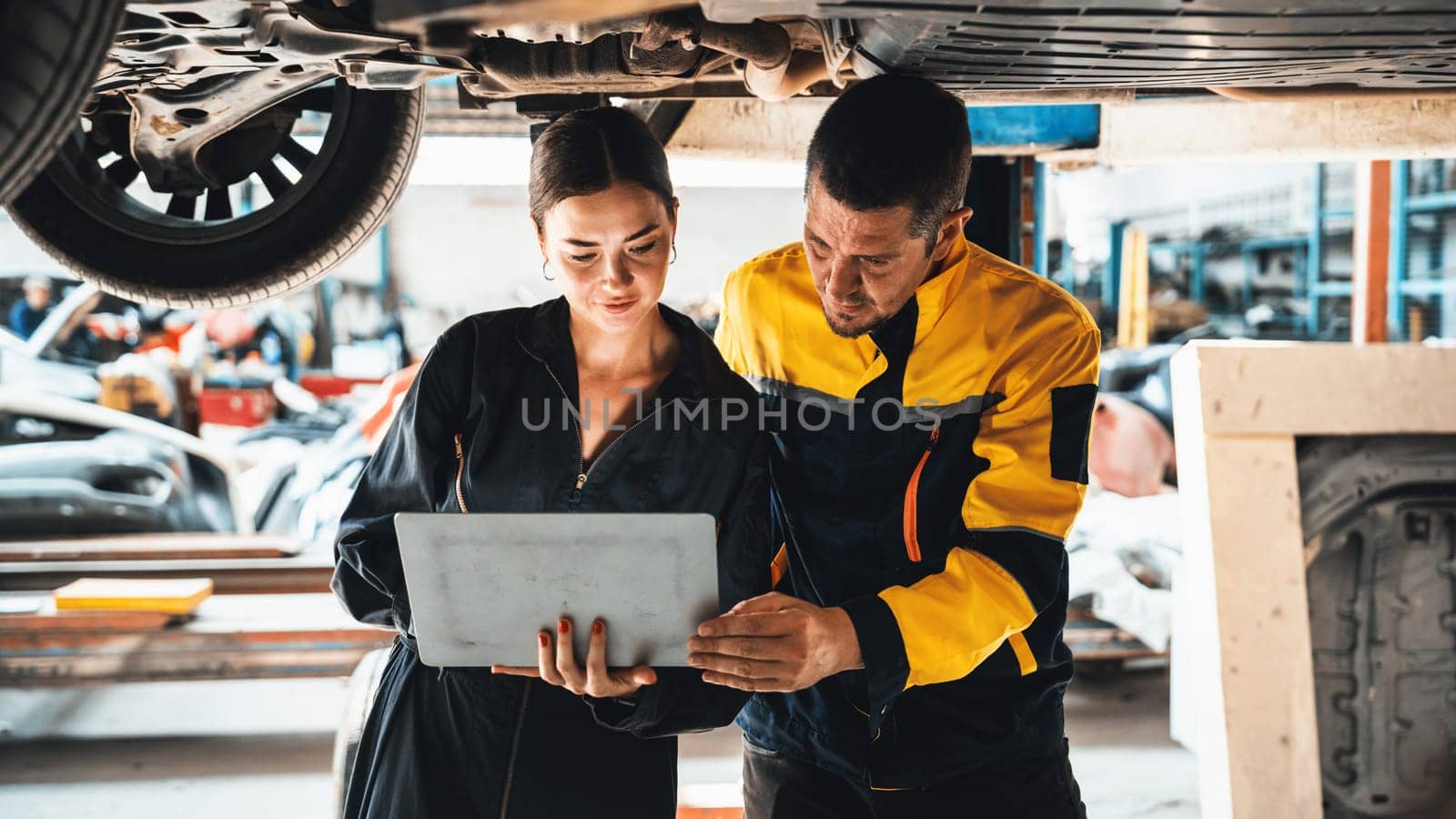 Two vehicle mechanic working together underneath lifted car, conduct car inspection with laptop. Automotive service technician in uniform carefully make diagnostic troubleshooting. Panorama Oxus