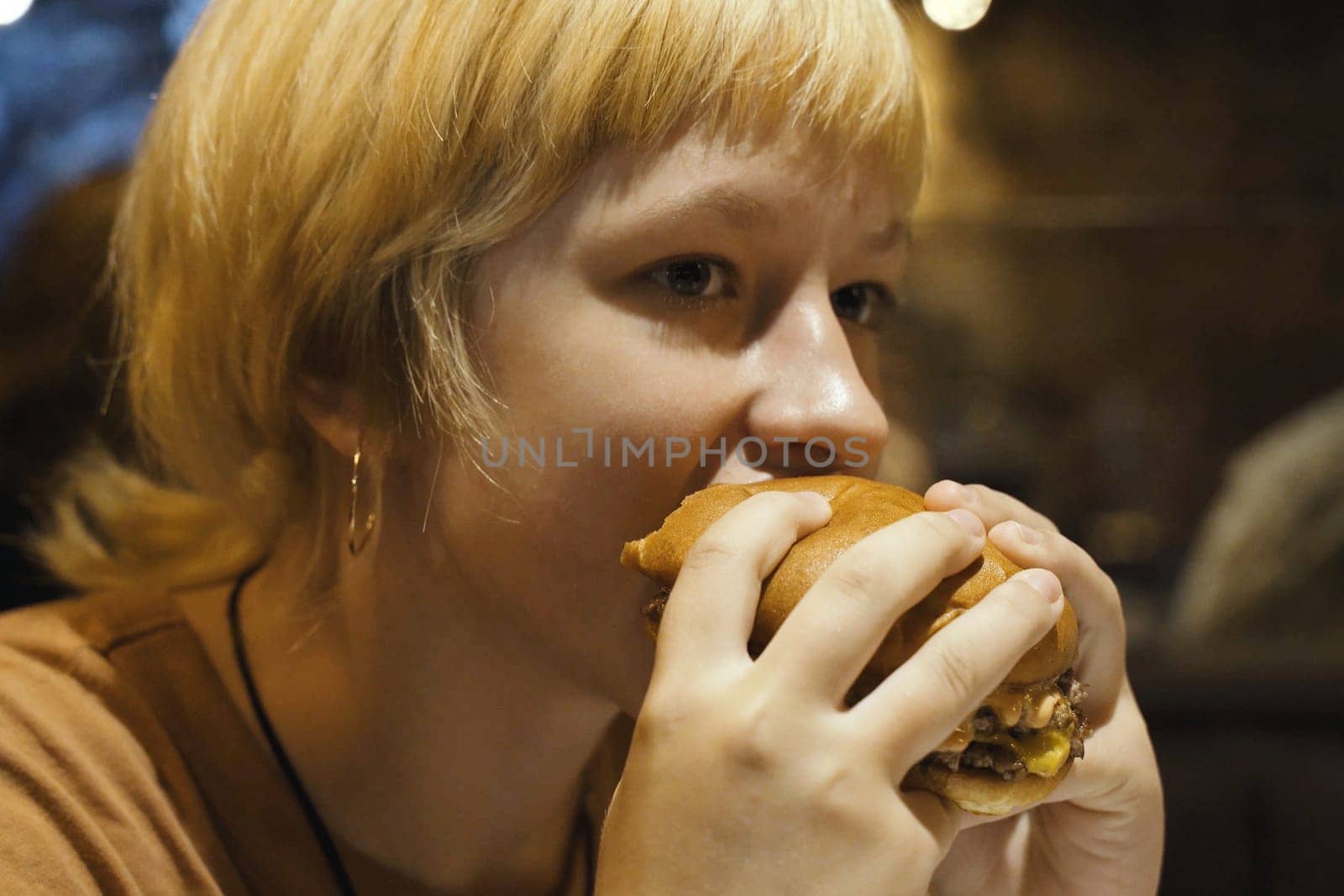 A teenager girl eats a hamburger in a restaurant, enjoying a delicious juicy burger. Close-up.