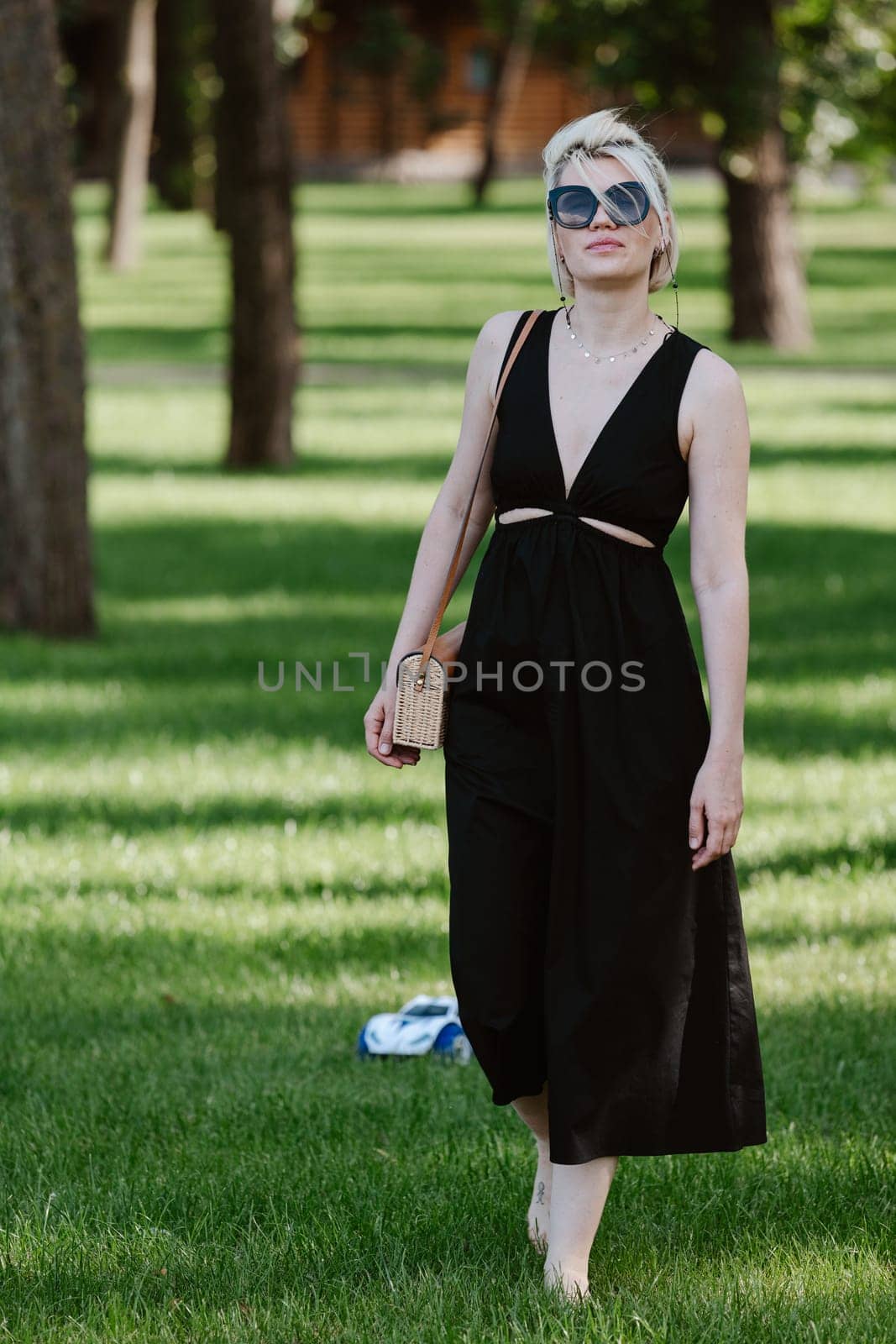 Young blond woman with short hair stands at full height in a park wearing sunglasses and black dress