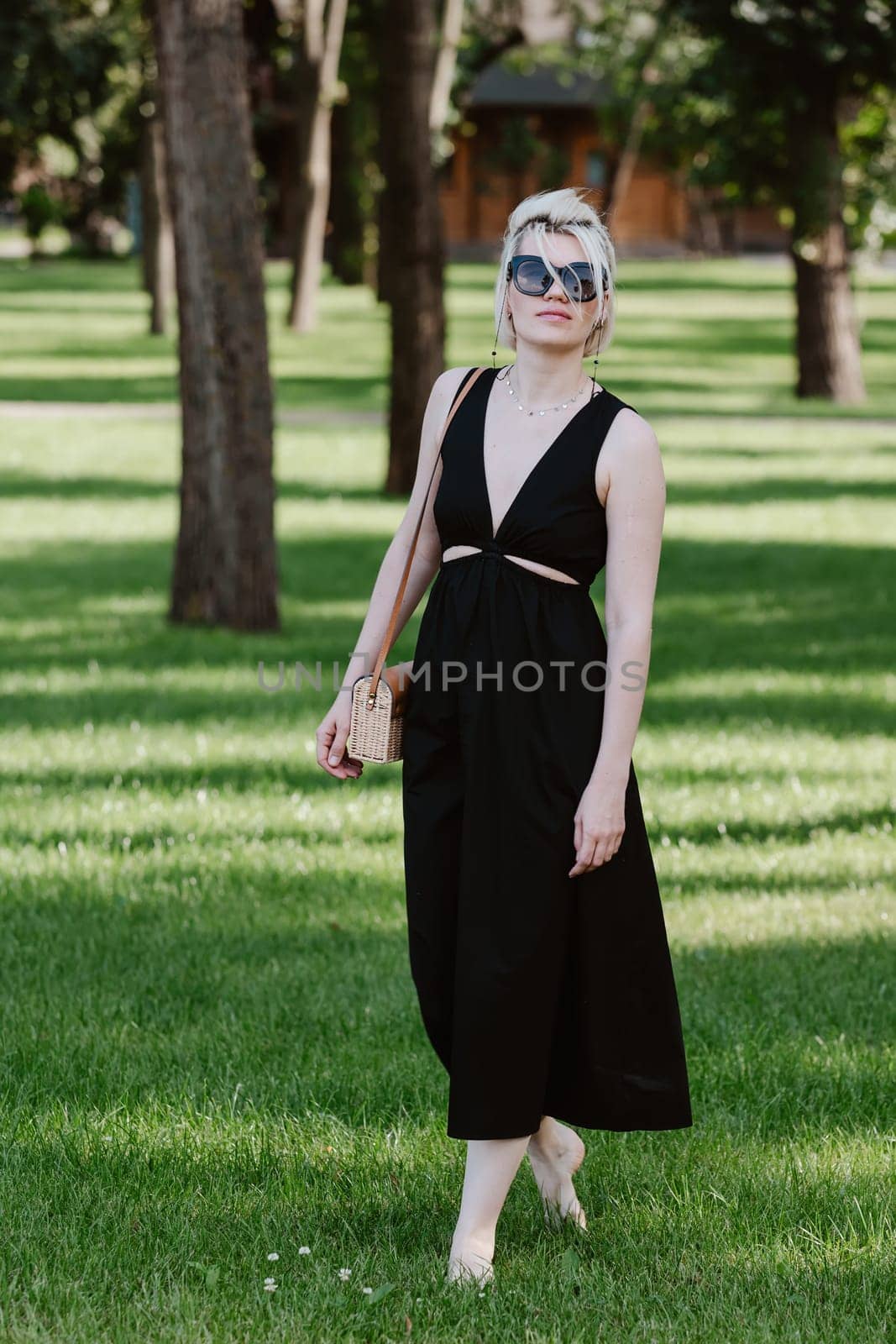 Young blond woman with short hair stands at full height in a park wearing sunglasses and black dress
